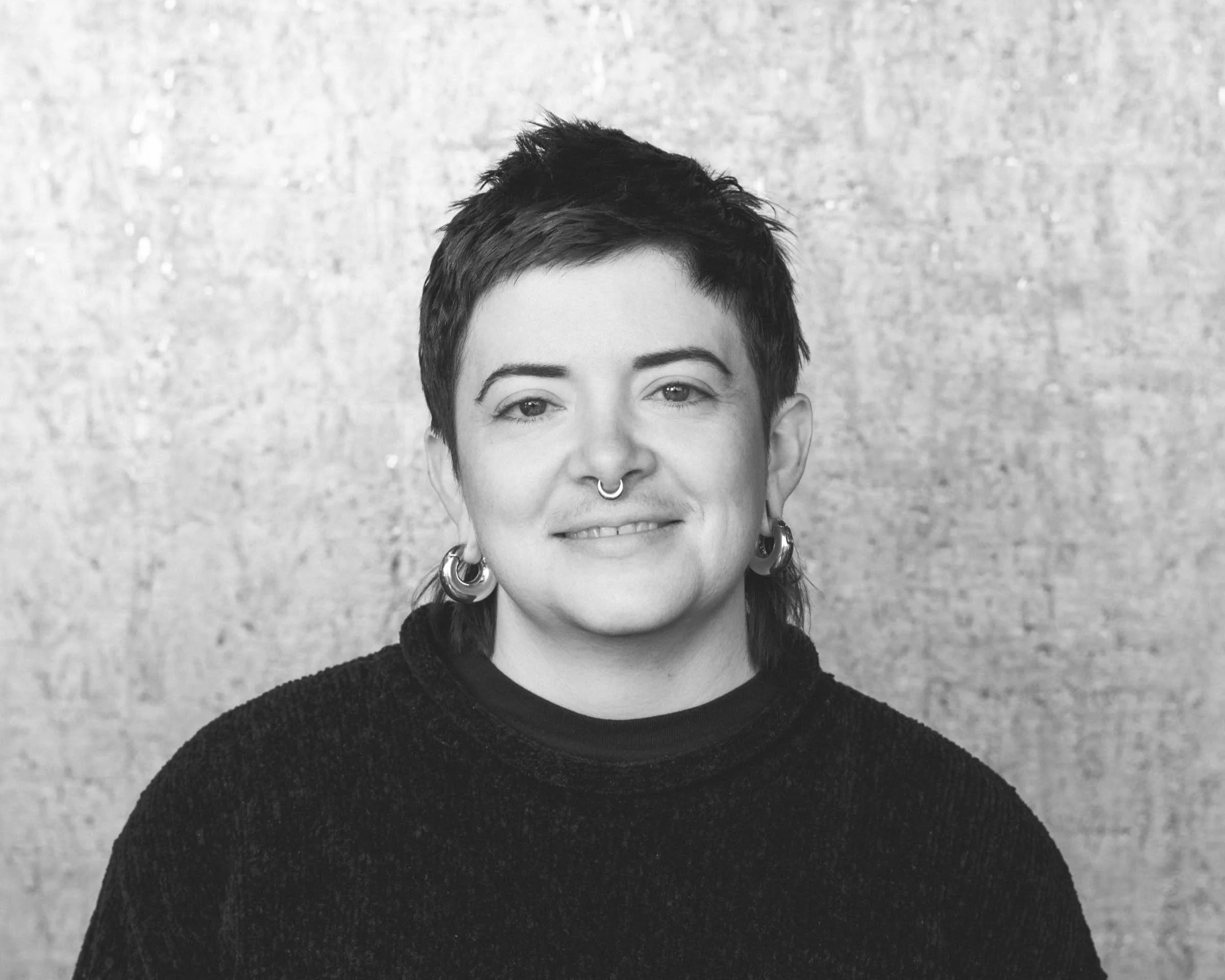 Black and white portrait of a woman with short, dark hair, multiple earrings, and a septum piercing, smiling slightly, wearing a dark sweater, in front of a textured light background.