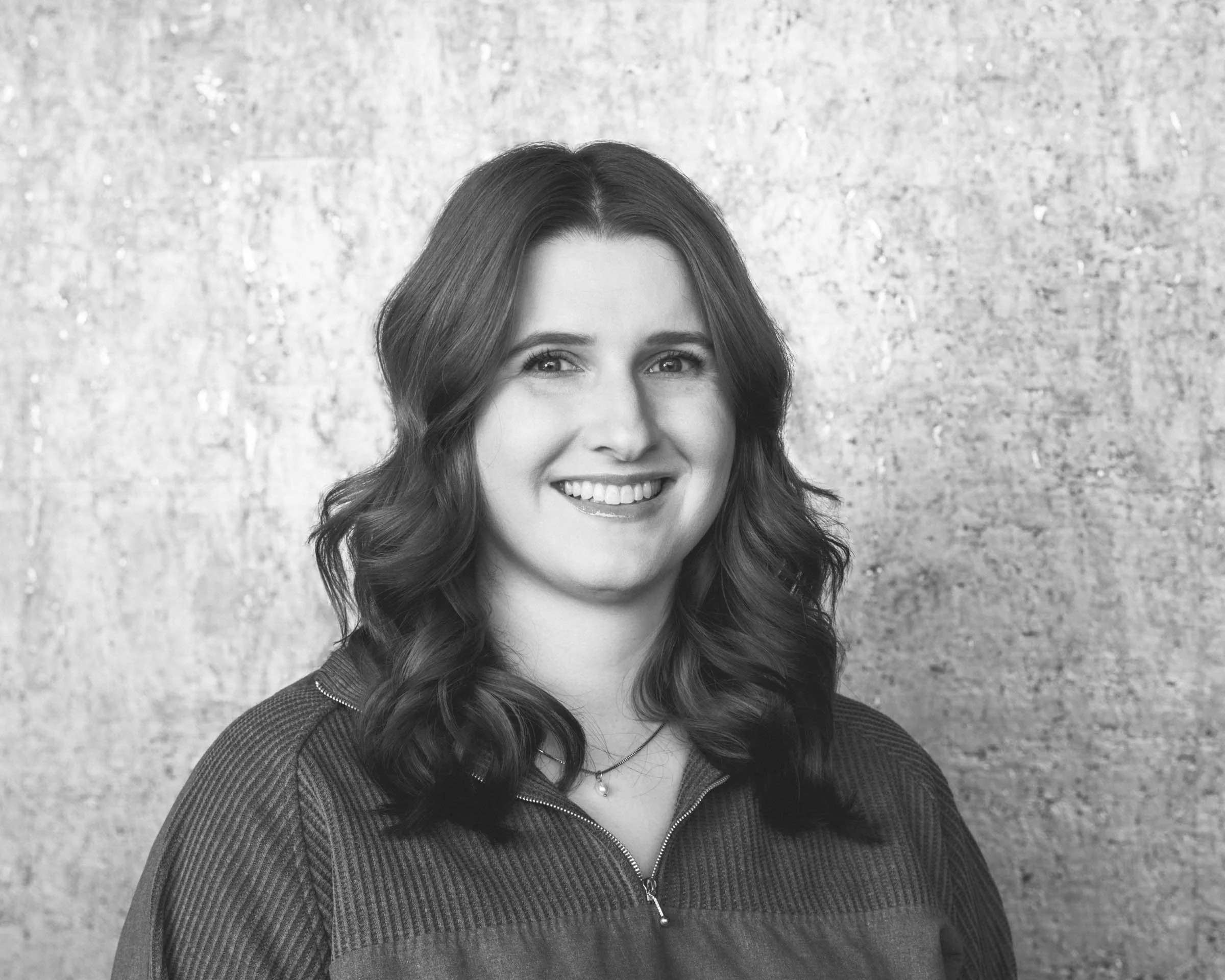 Black and white portrait of a smiling woman with wavy hair, wearing a zip-up jacket, standing against a textured background.