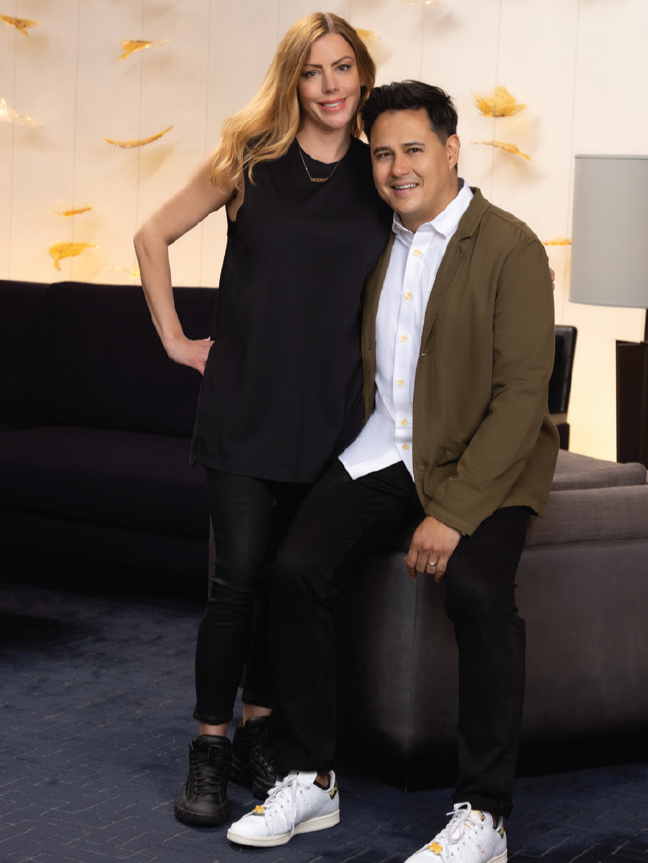 A woman with long blonde hair and a man with dark hair and a light skin tone, smiling together in a modern living room. The woman wears a sleeveless black top and black pants, and the man wears a white shirt, olive-green jacket, and black pants.