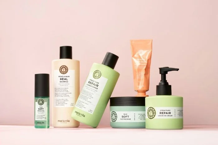 A collection of hair and skincare products on a pastel pink background. The products include shampoo, conditioner, leave-in cream, and other treatments in bottles and jars, mostly in soft green, cream, and peach colors.