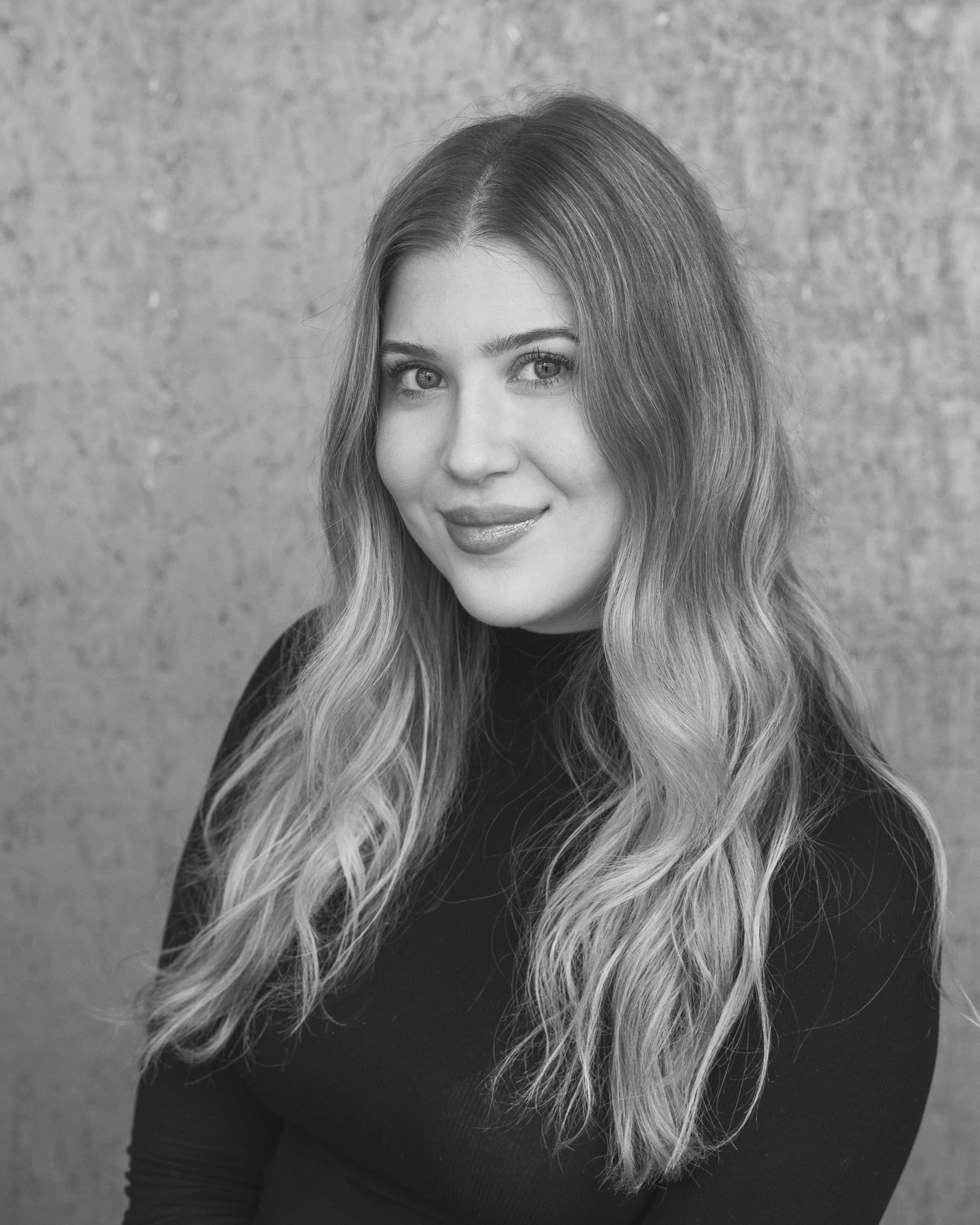 Black and white portrait of a young woman with long, wavy hair, smiling softly, wearing a dark top, standing against a textured wall.