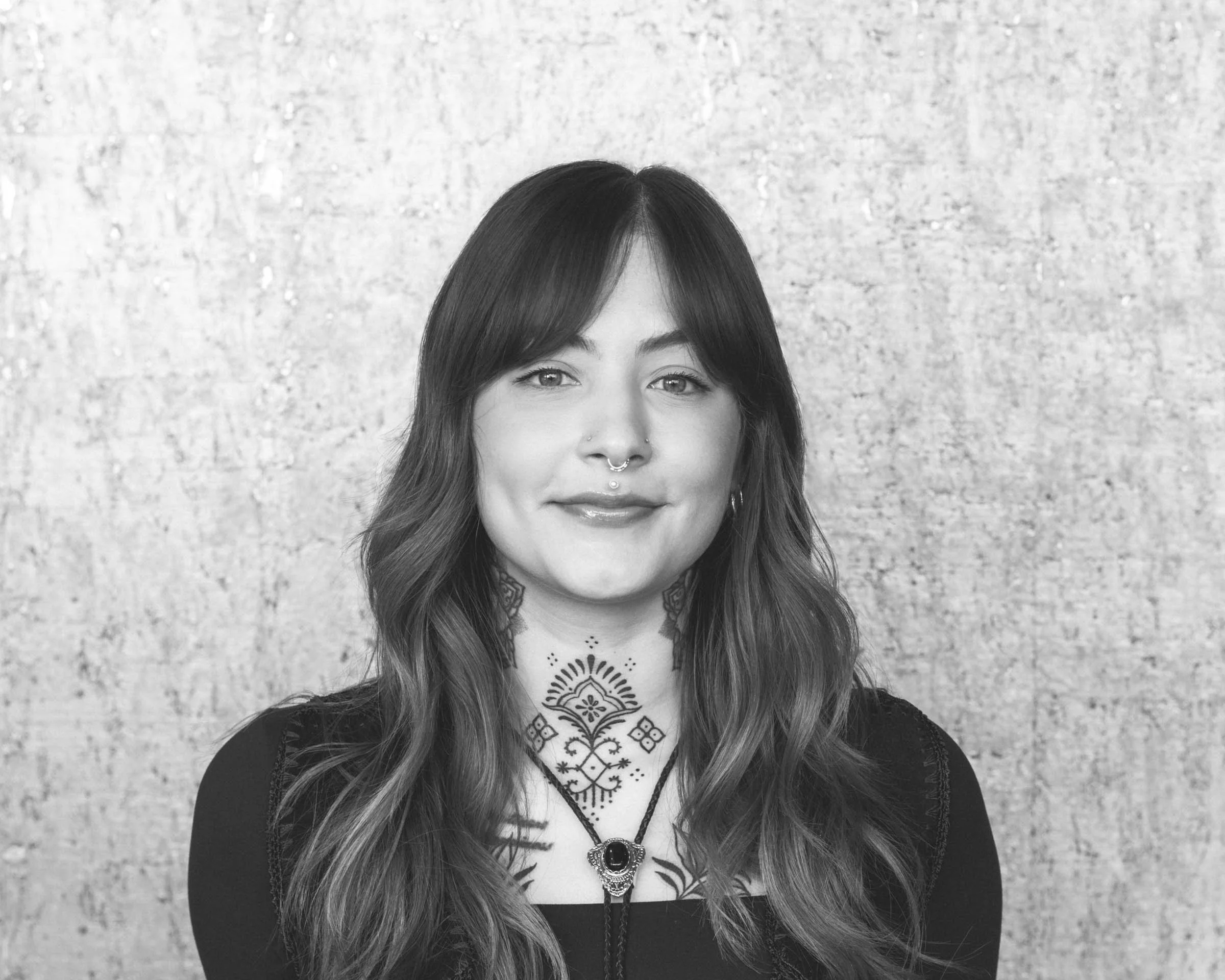Black and white portrait of a woman with long wavy hair, sporting tattoos, piercings, and wearing a necklace, standing against a textured wall.