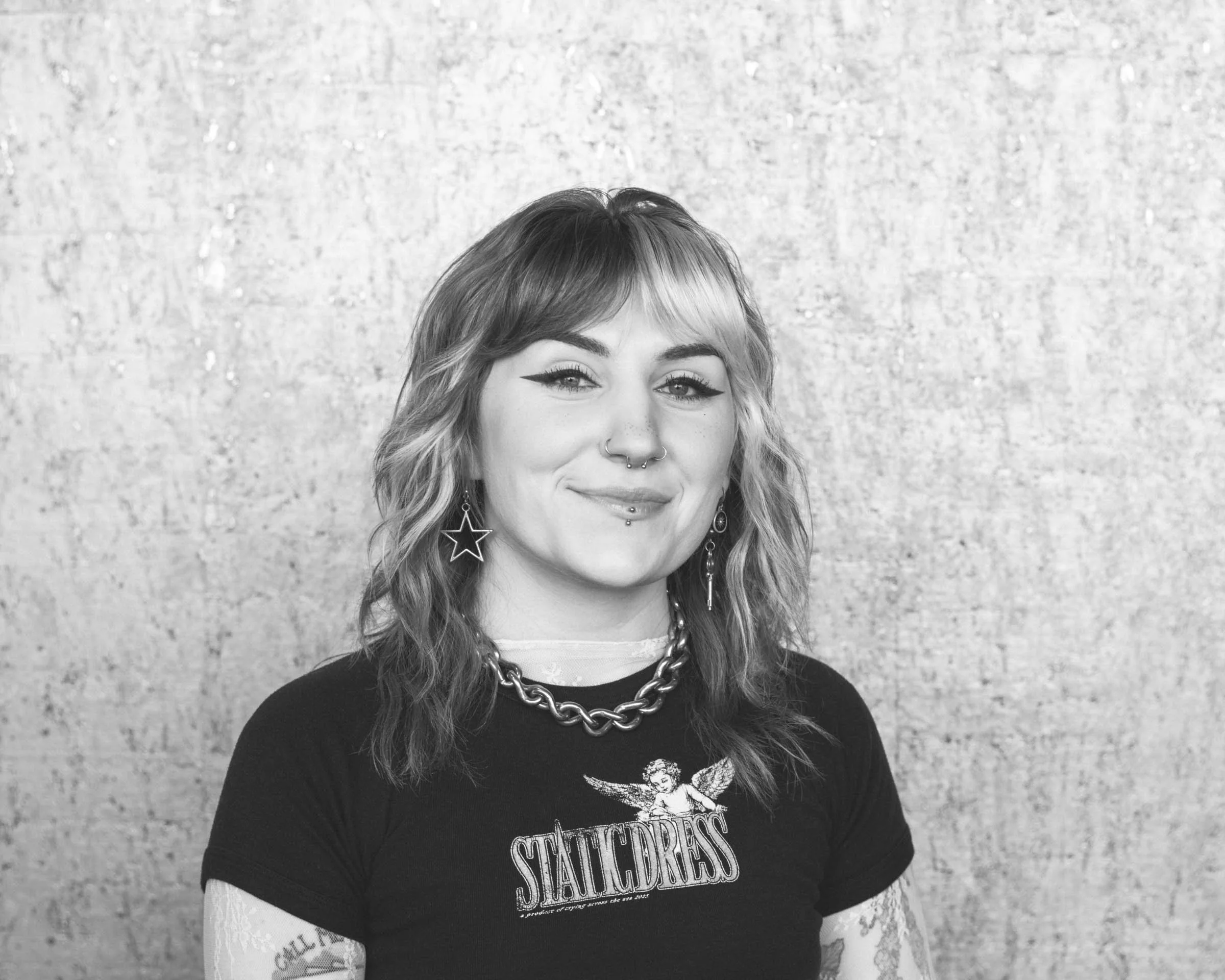 A young woman with wavy hair, smiling, wearing a black t-shirt with a graphic, star-shaped earrings, layered necklaces, and tattoos on her arms, standing against a textured background.
