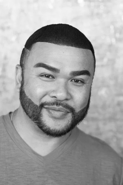 Black and white portrait of a man with short hair, full beard, and a slight smile, wearing a V-neck shirt, against a blurred background.