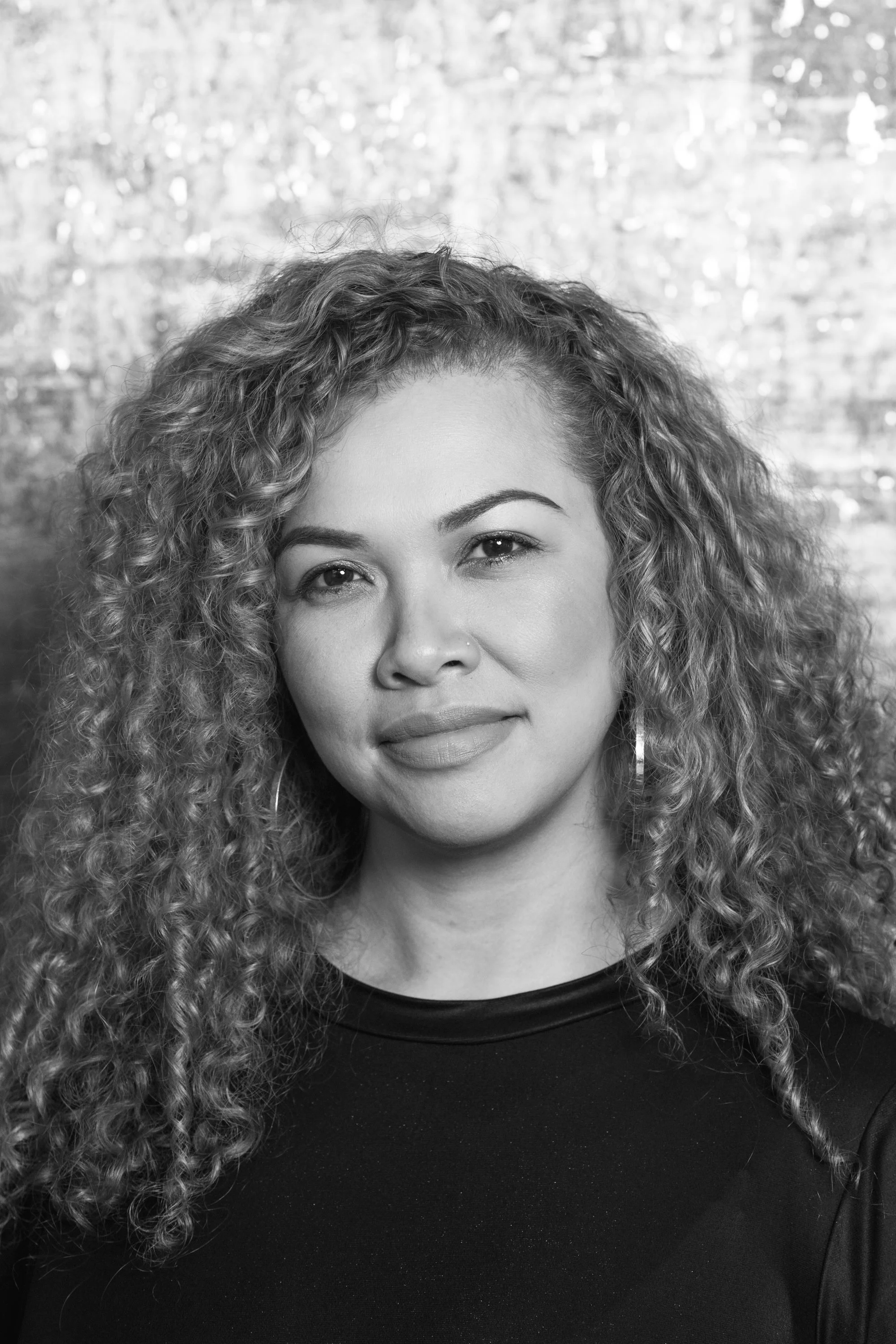 Black and white portrait of a woman with curly hair, wearing a dark top and hoop earrings, looking at the camera with a slight smile.