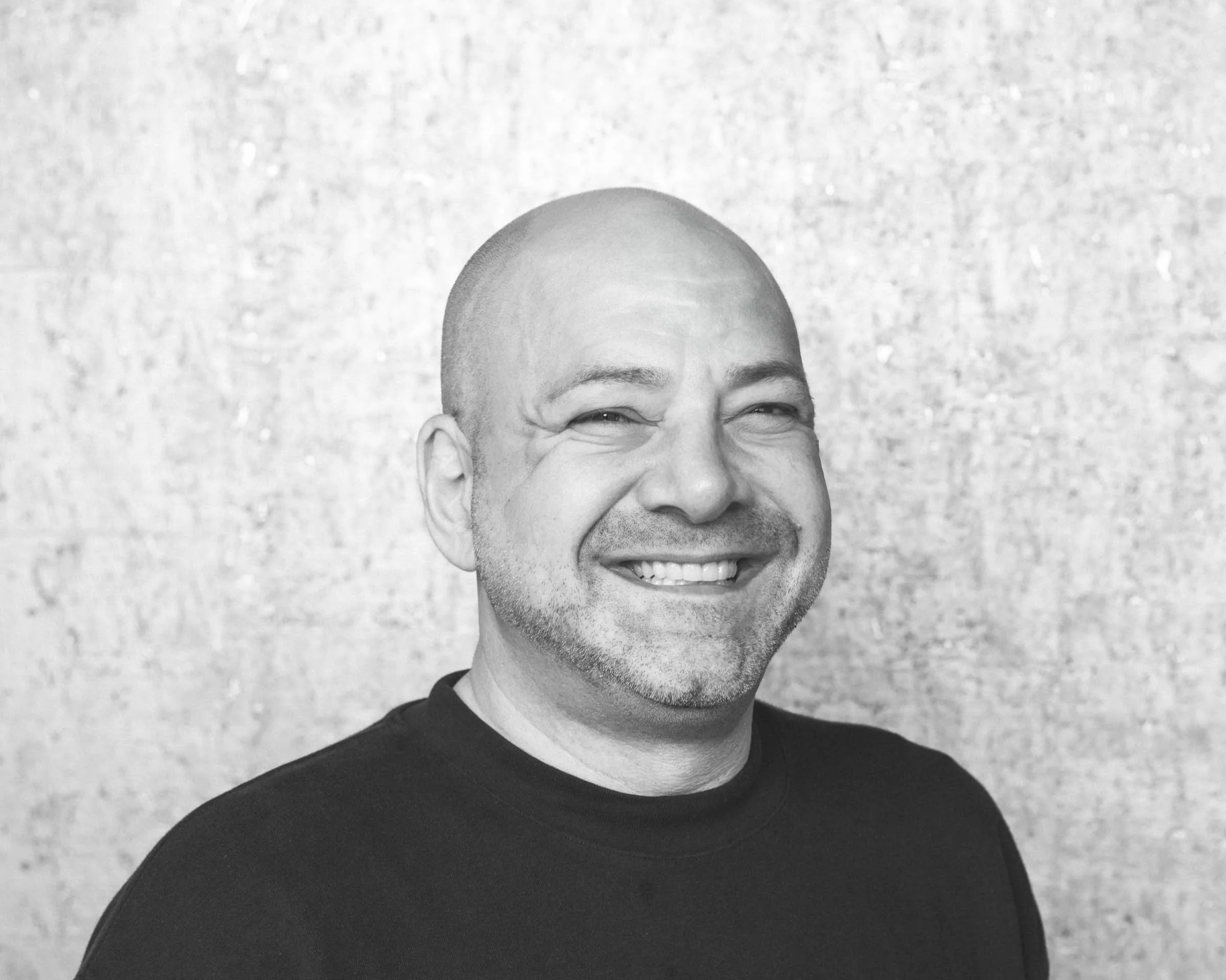Black and white photo of a smiling, bald man with a beard, wearing a dark shirt, standing in front of a textured wall.