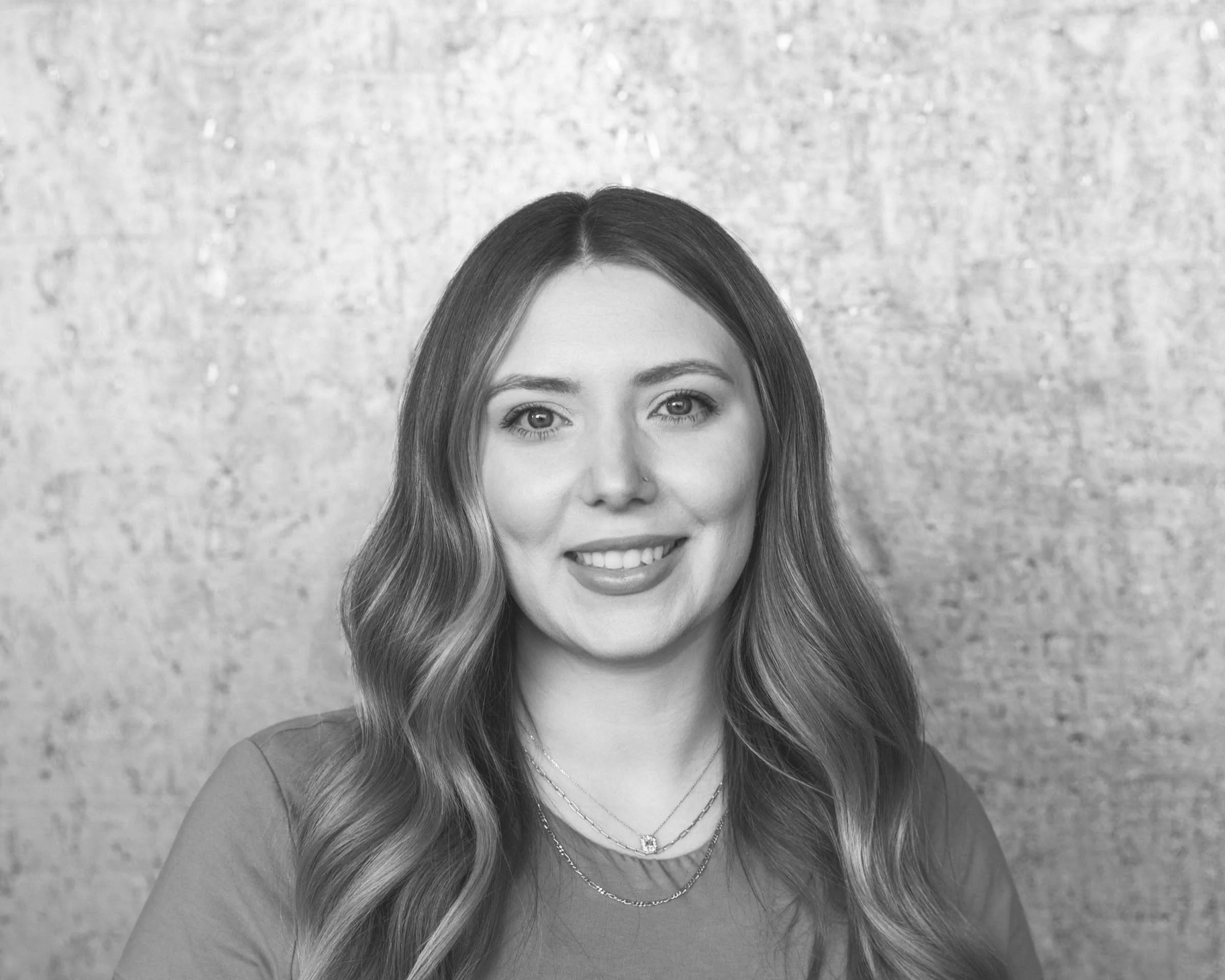 A grayscale portrait of a woman with wavy hair, smiling, wearing layered necklaces, against a textured wall background.