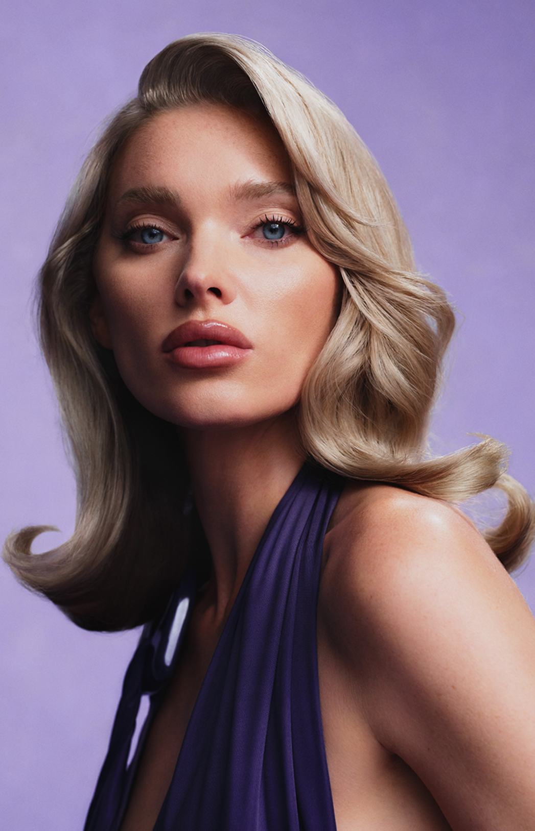 A woman with blonde wavy hair and blue eyes wearing a purple sleeveless dress against a purple background.