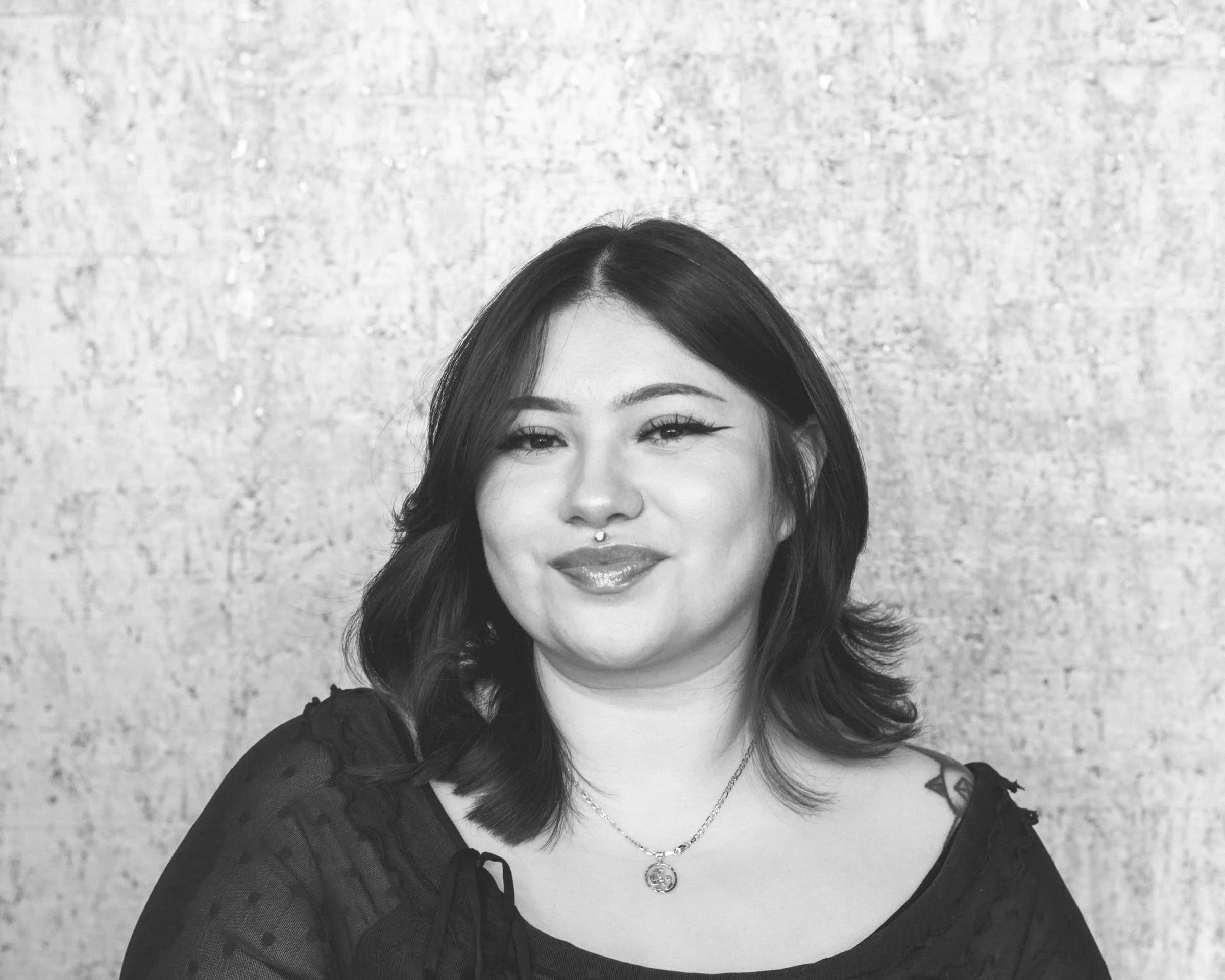 A woman with shoulder-length dark hair, winking and smiling, wearing a necklace and a black top with sheer sleeves, standing against a textured wall background.