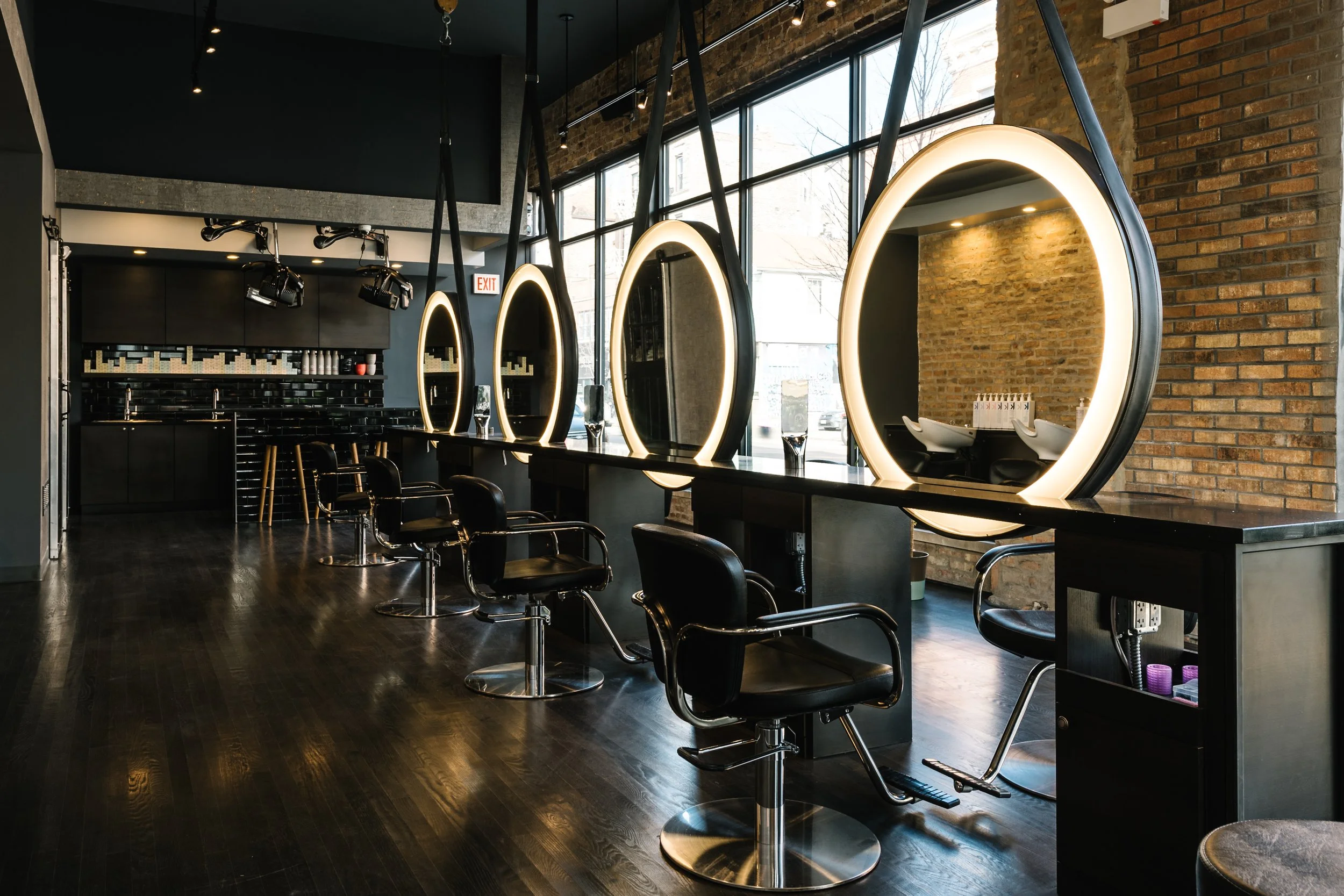Empty hair styling station with black chairs, large illuminated oval mirrors, black countertops, and a brick wall inside a modern salon.