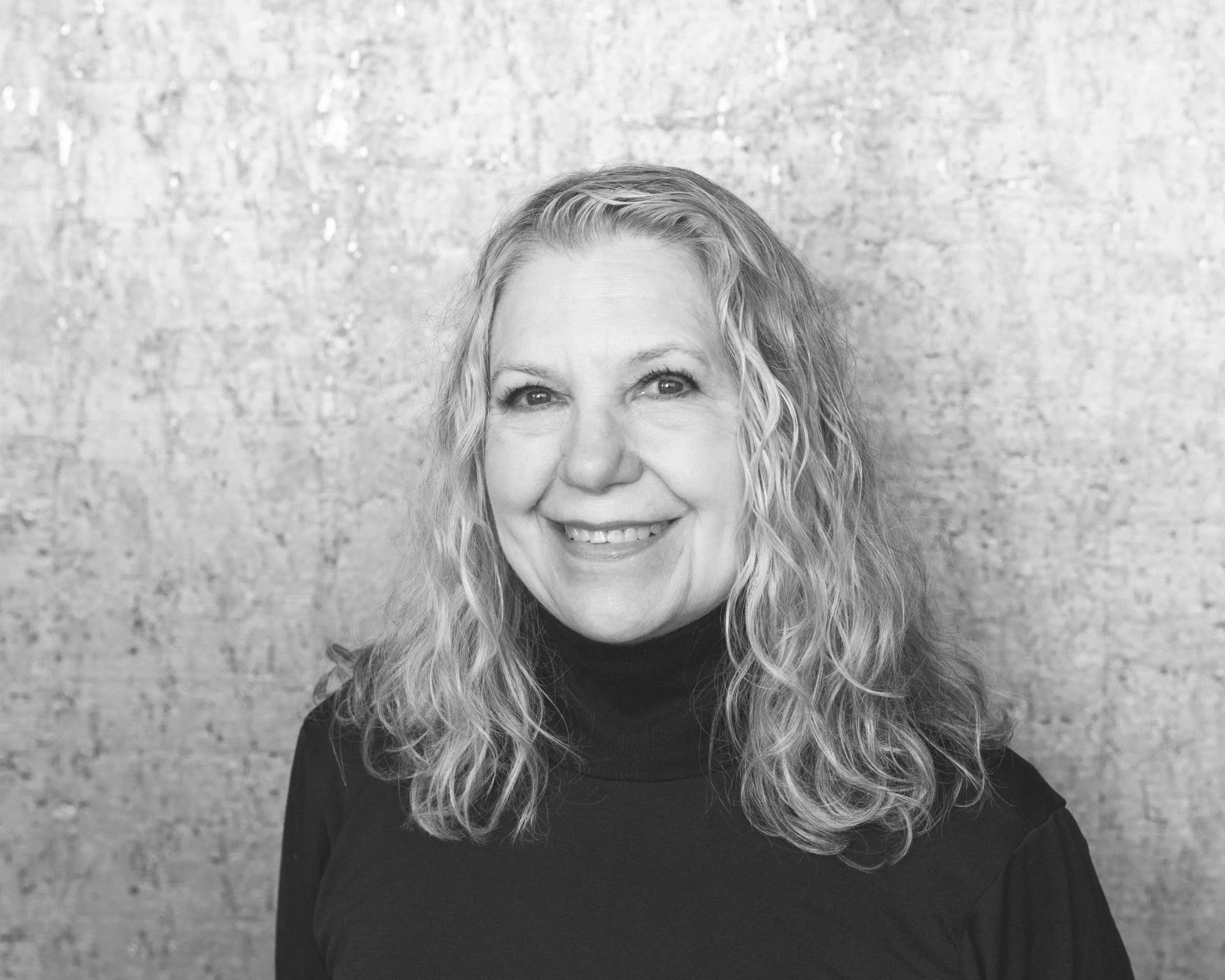 Black-and-white portrait of a smiling middle-aged woman with curly hair wearing a black turtleneck, standing against a textured background.