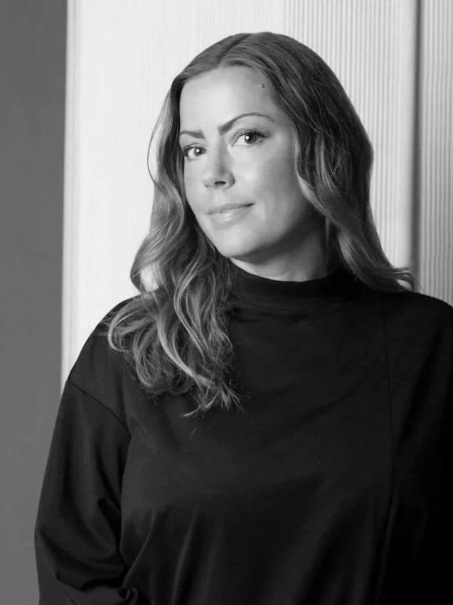 Black and white photo of a woman with wavy hair, wearing a black top, standing indoors beside a wall with vertical paneling, looking at the camera with a slight smile.
