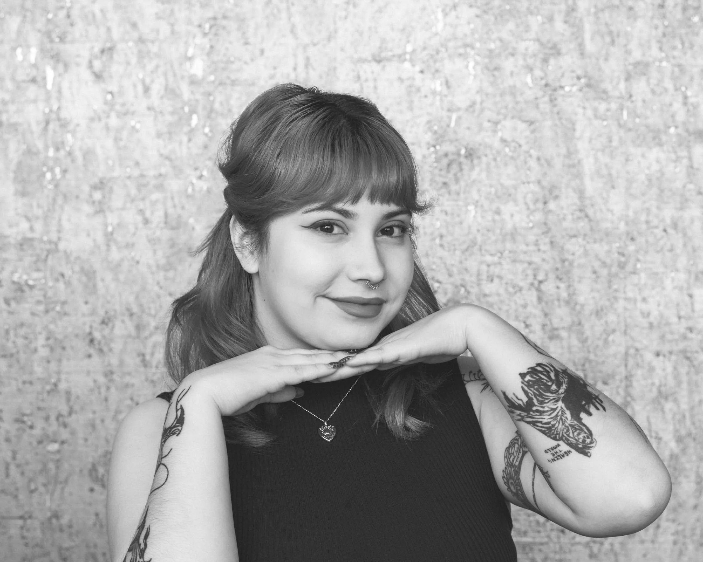 A young woman with tattoos on her arms and a septum piercing, smiling with her hands under her chin, posing in front of a textured wall.
