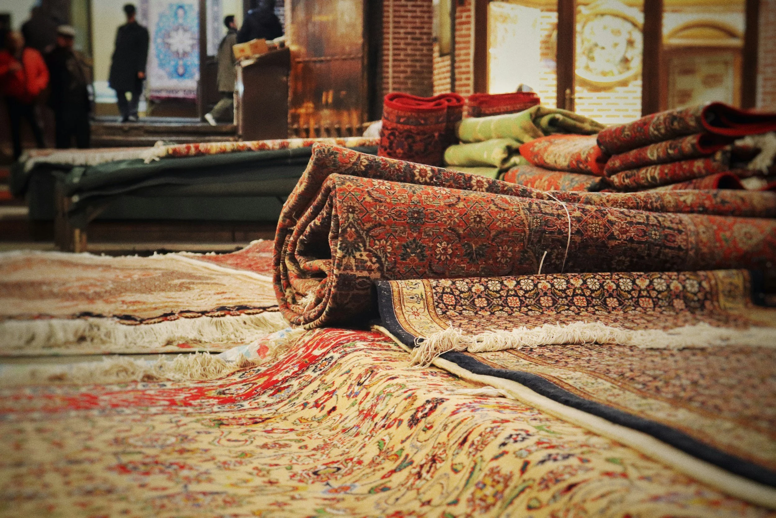Stacks of colorful, patterned rugs displayed on the floor inside an indoor market or store, with some rolled up and others spread out.