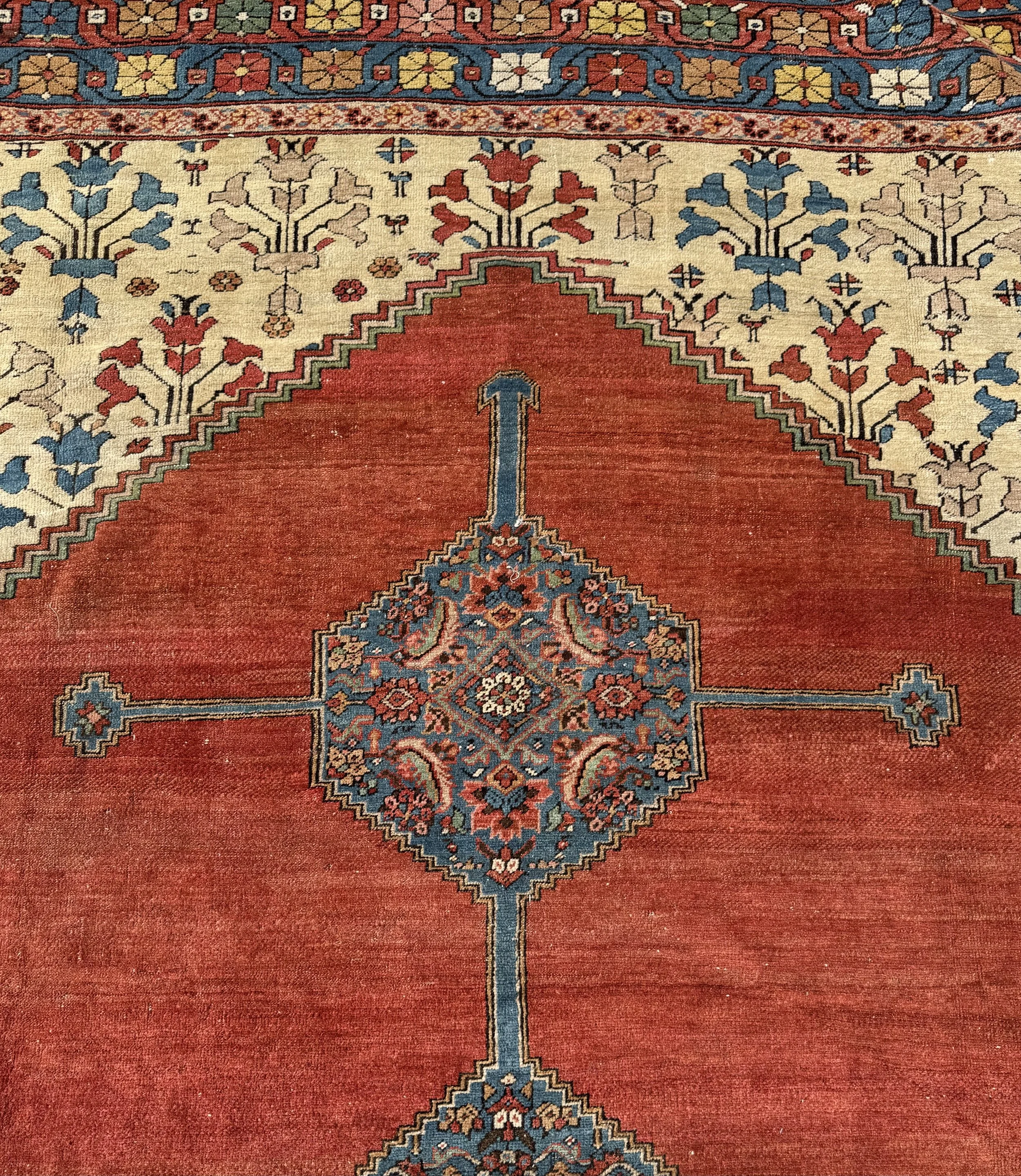 Close-up of a traditional patterned rug with a red background, featuring a central blue and red medallion, surrounded by intricate floral designs and borders in shades of cream, yellow, blue, and red.