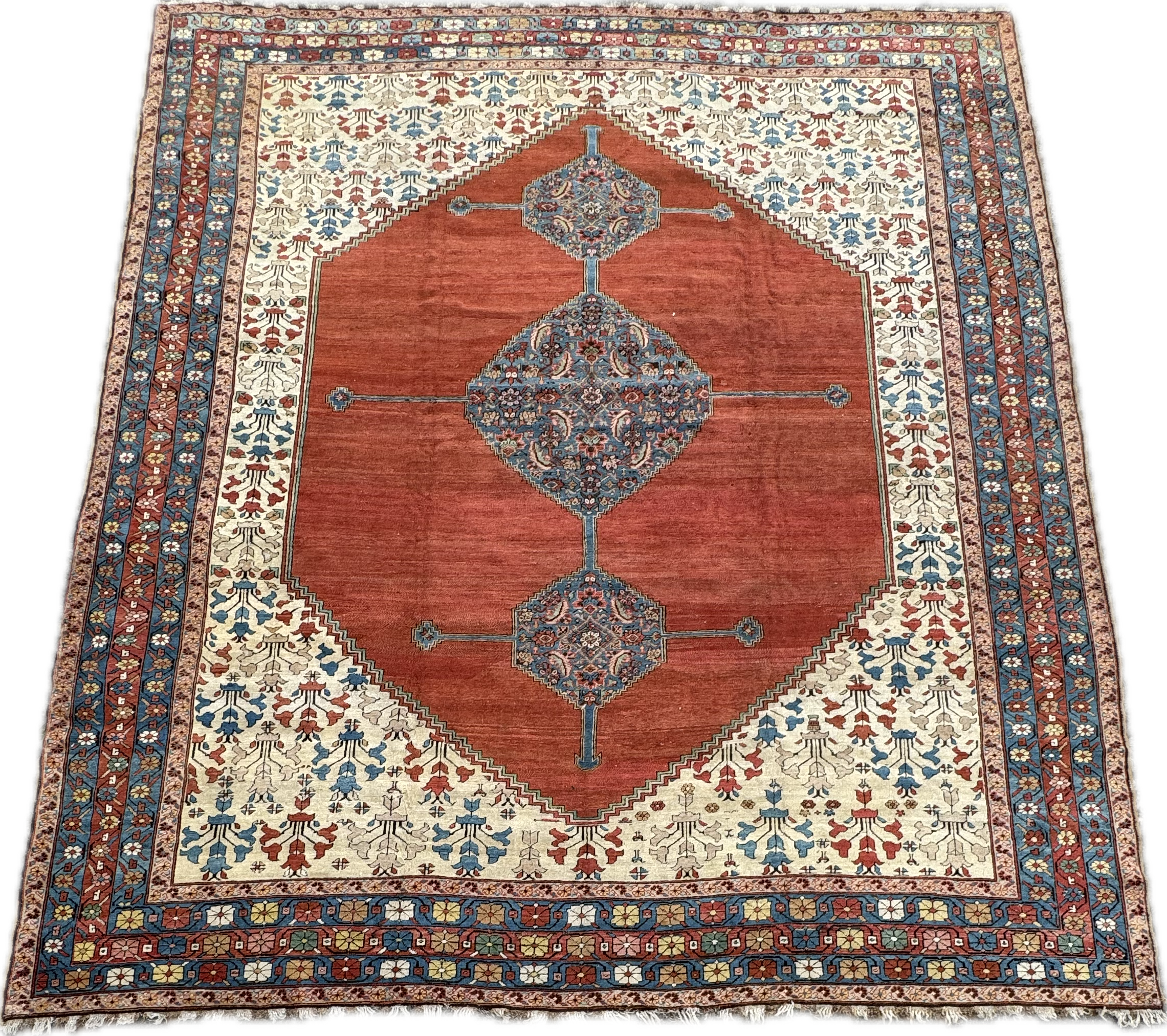 A traditional rectangular area rug with a central red field, ornate floral medallions, and multiple borders featuring intricate floral and geometric patterns in cream, blue, red, and tan colors.