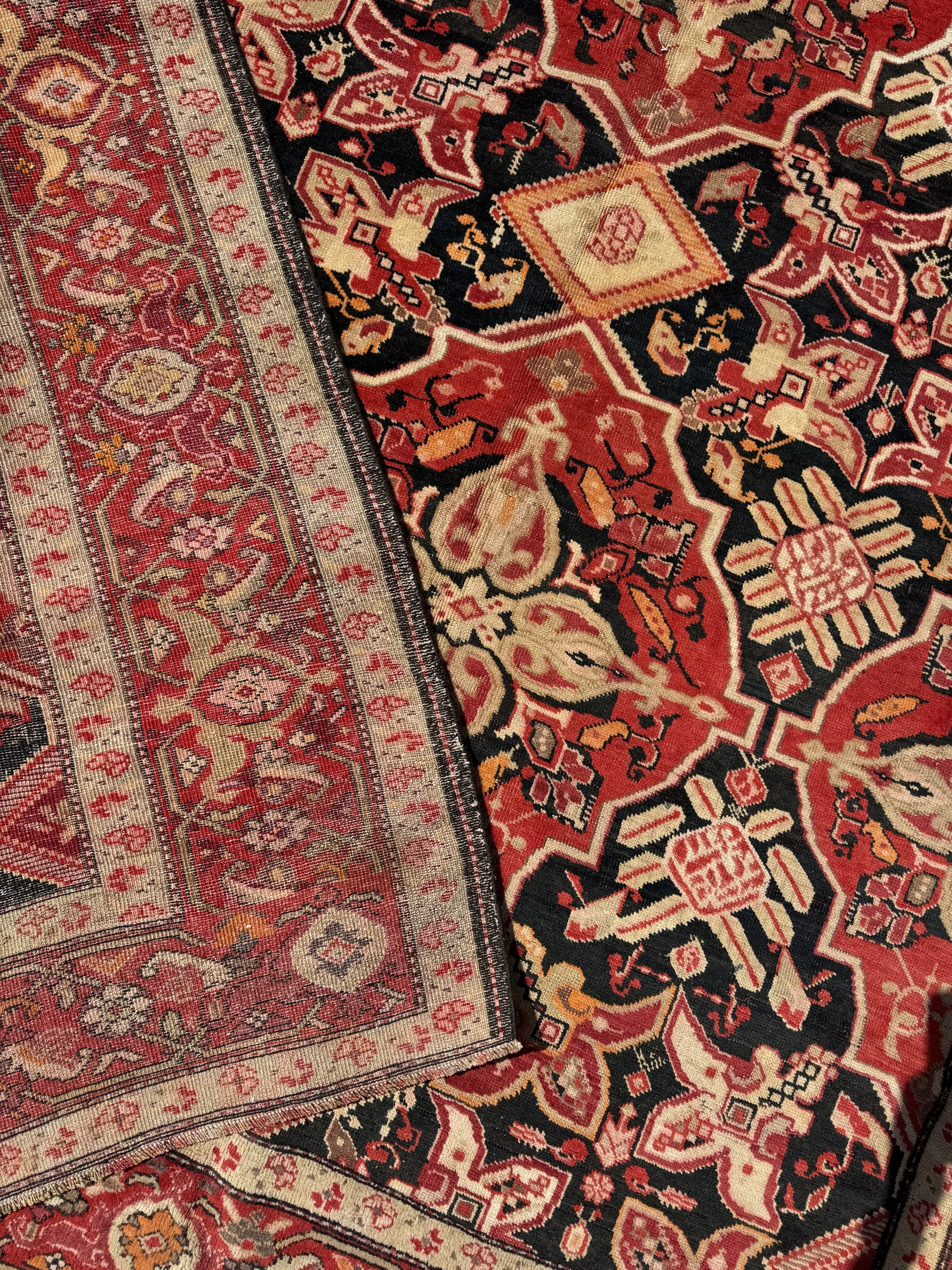 Close-up of two overlapping traditional Persian rugs with intricate floral and geometric patterns in red, black, beige, and orange colors.