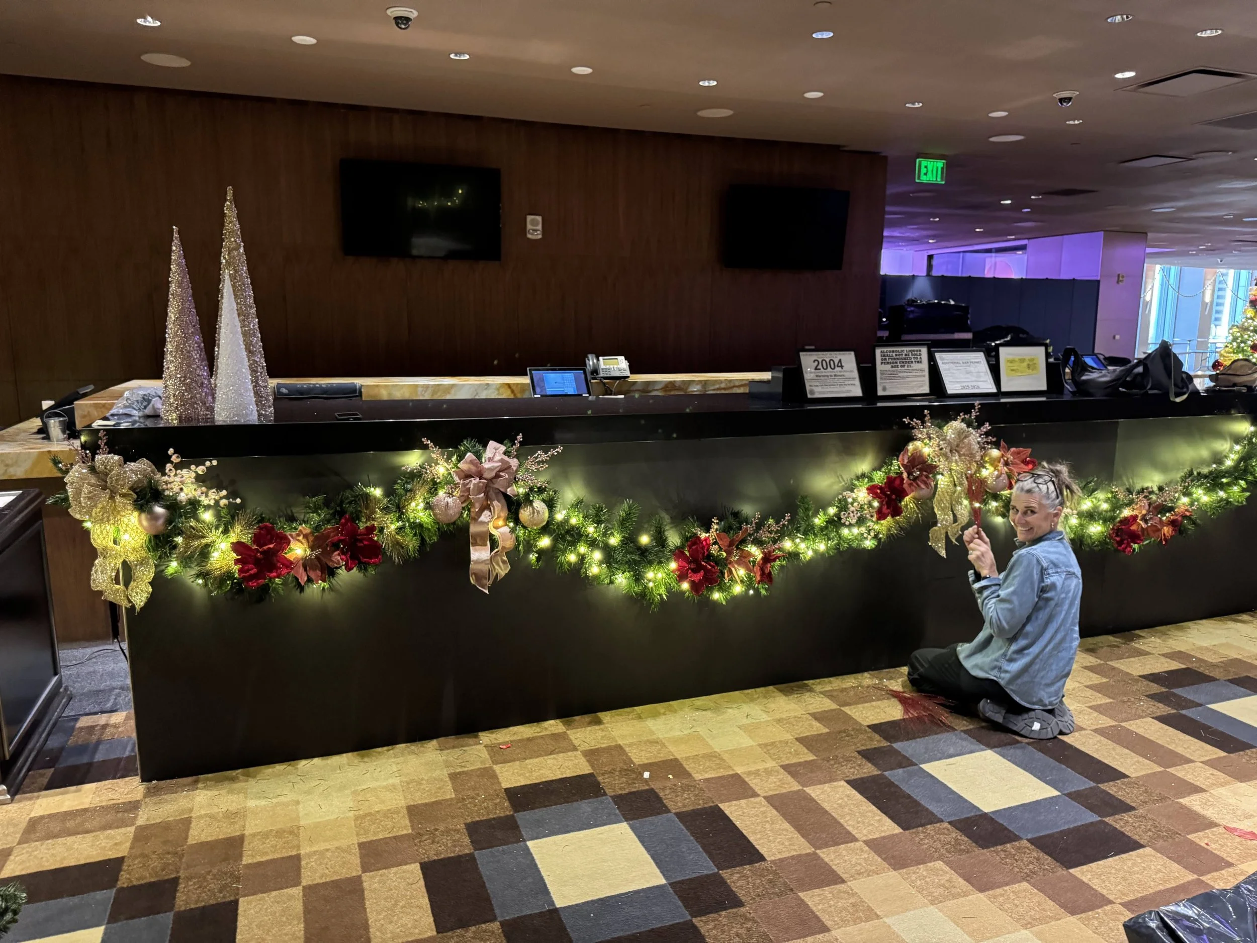A woman is kneeling in front of a decorated counter with Christmas ornaments and greenery, holding a candy cane and smiling at the camera in a hotel lobby with Christmas decor and holiday lights.