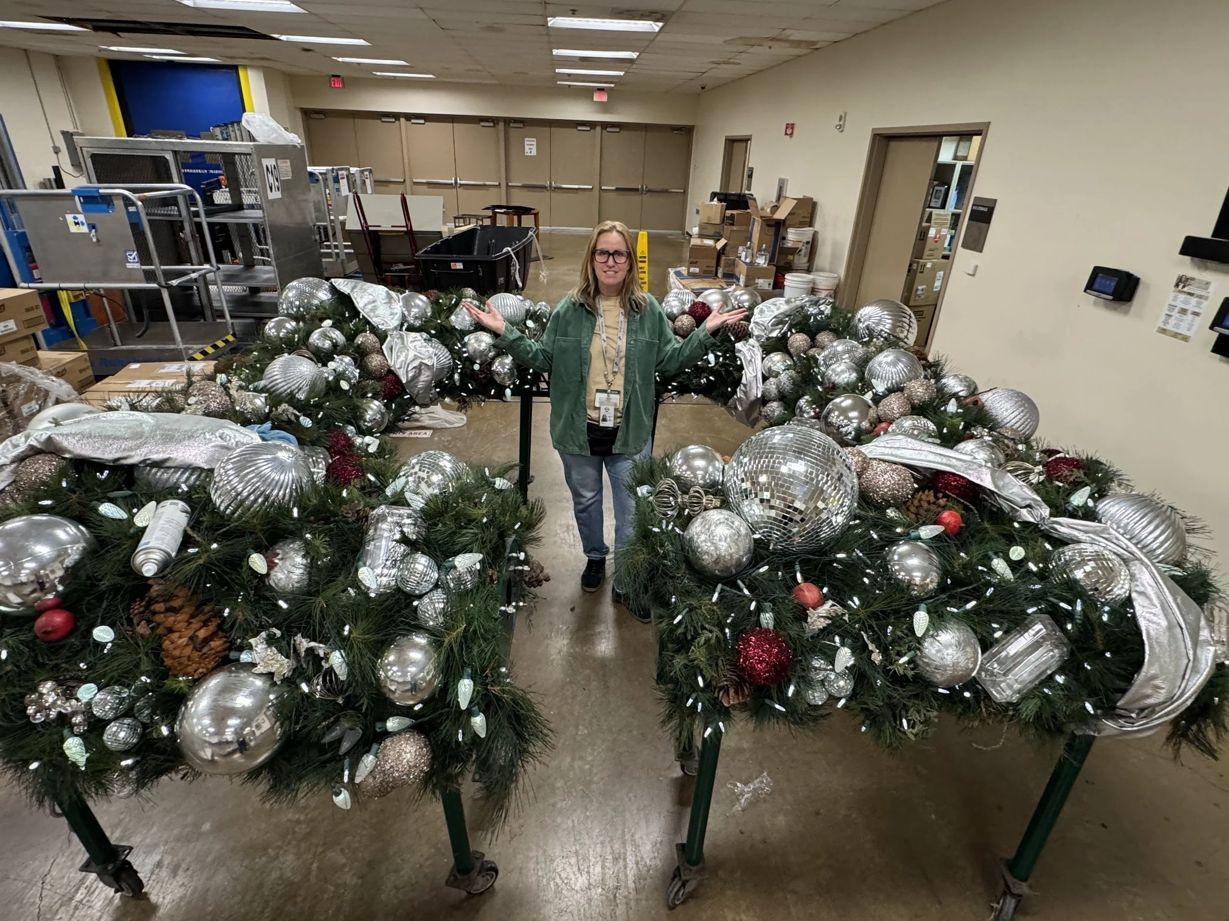 A woman standing behind a festive Christmas display of silver, red, and glittery ornaments and decorations in a warehouse or store setting.