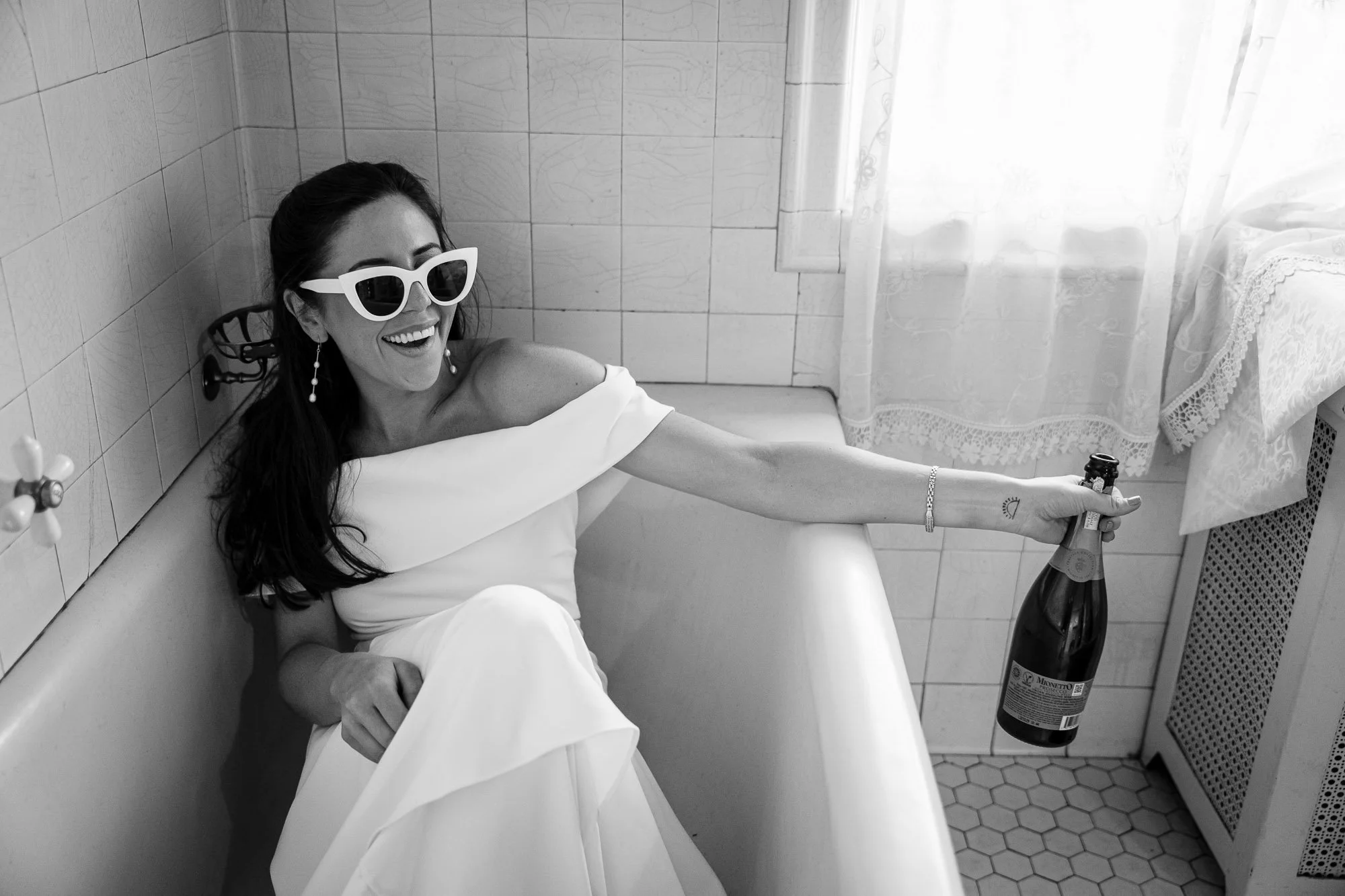 A black and white image of a bride chillin' in a classic bathtub during a break on her Chicago Summer wedding day, wearing sunglasses and holding a bottle of champagne.
