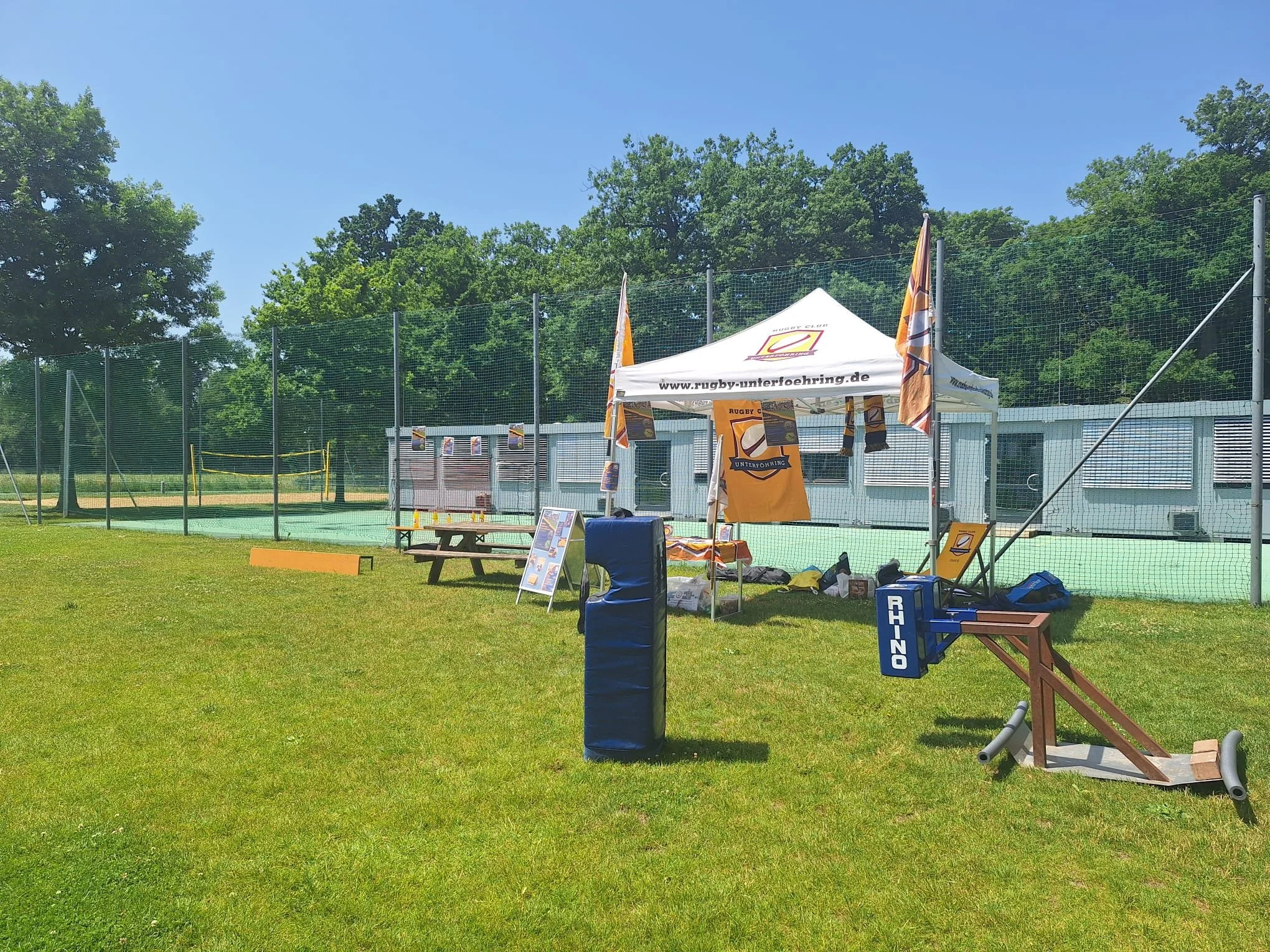 Rugby equipment set up outdoors on a grassy field with a white tent, flags, and benches, near a sports court enclosed by a tall fence.