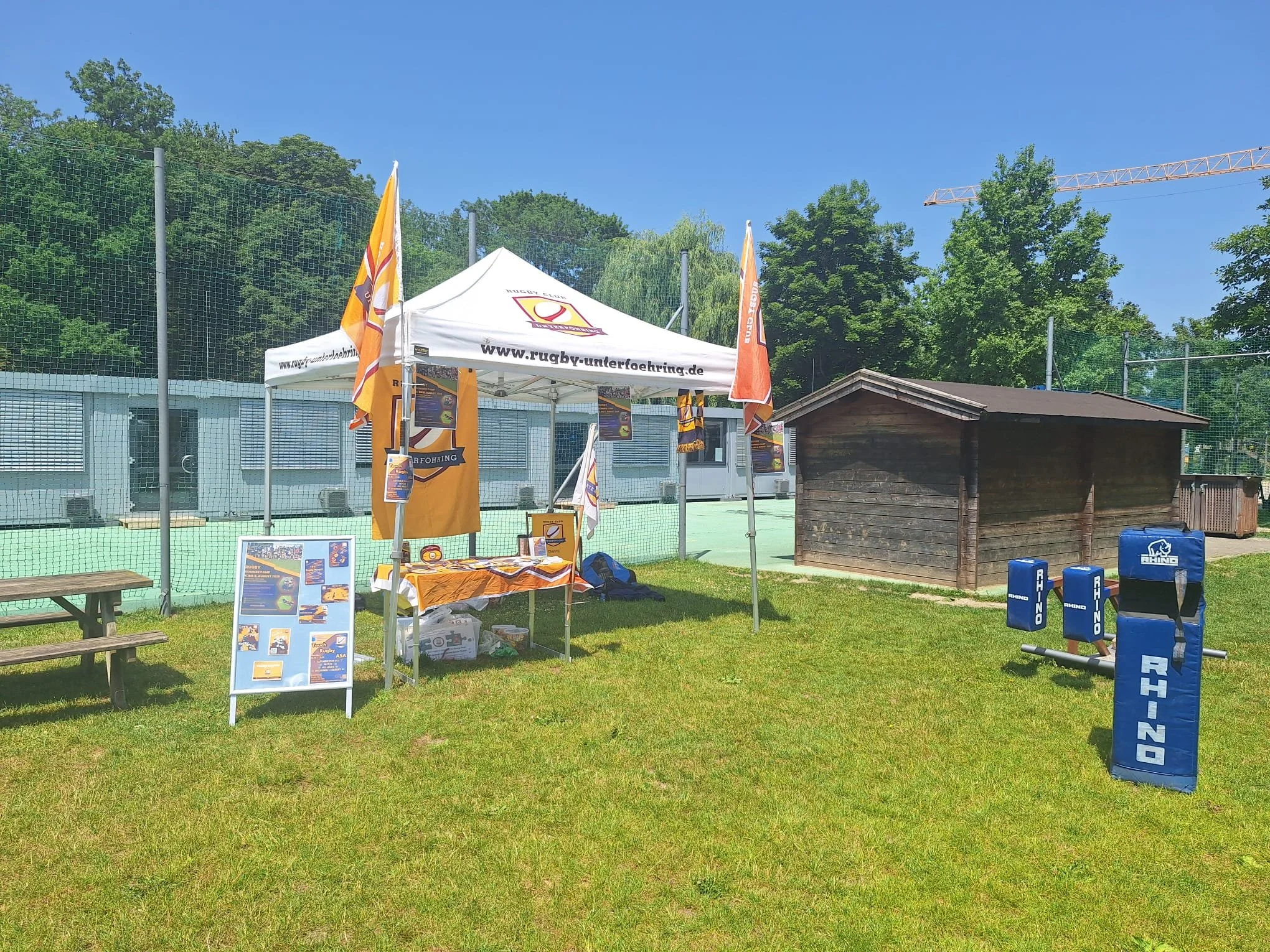 Rugby promotional booth with orange and white flags, posters, and tables set up outdoors near a sports field, with a small wooden shed and exercise equipment nearby.