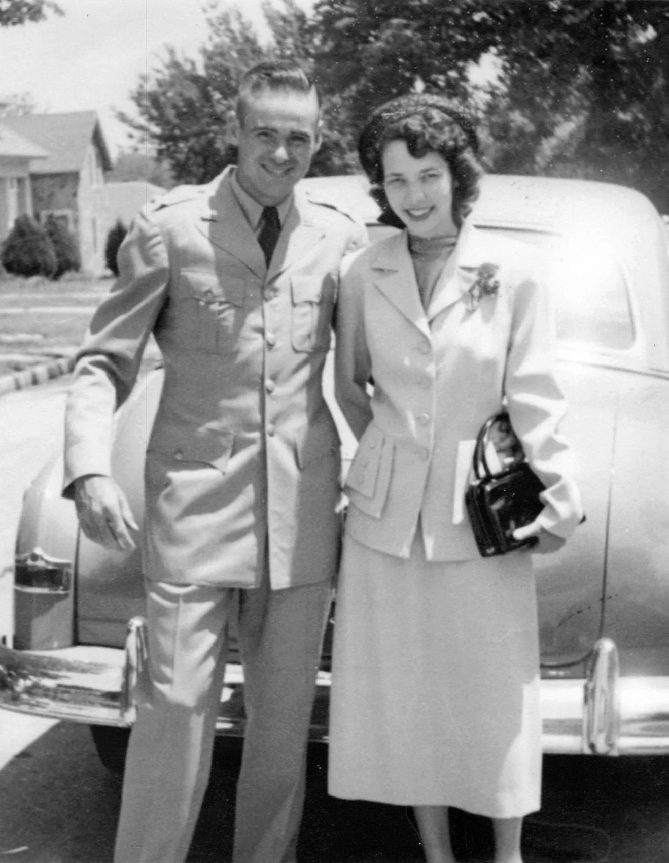 A black-and-white photo of a man in a military uniform and a woman in a formal outfit standing outdoors in front of a vintage car, with houses and trees in the background.