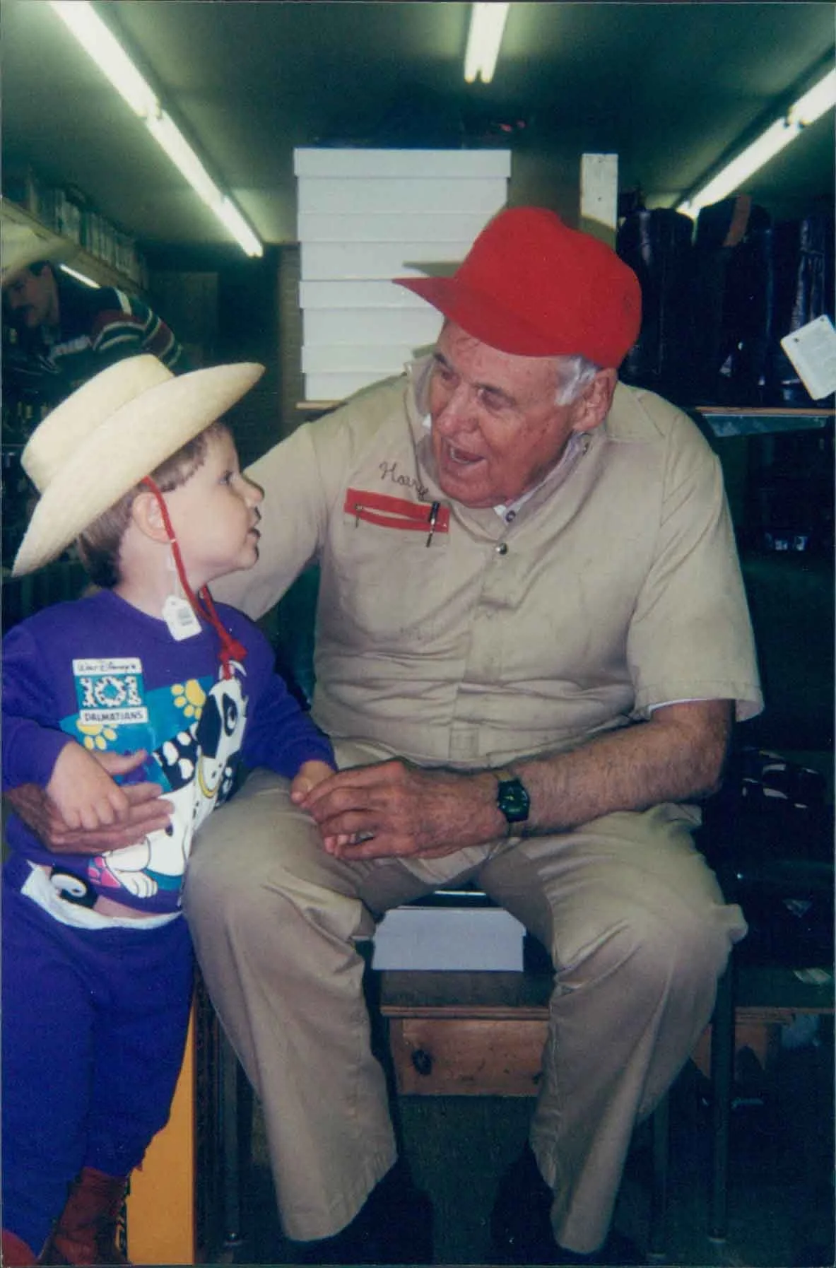 An older man with white hair, wearing a red cap and beige outfit, sitting on a chair and talking to a young boy in a straw hat and colorful shirt, inside a store.