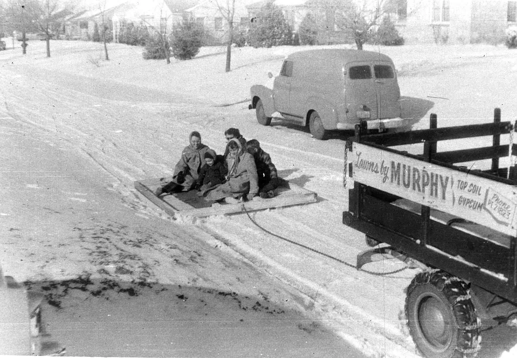 A group of people sitting on a sled in a snowy landscape with a vintage van and a truck with a sign that reads "Tourney by Murphy" in the background.