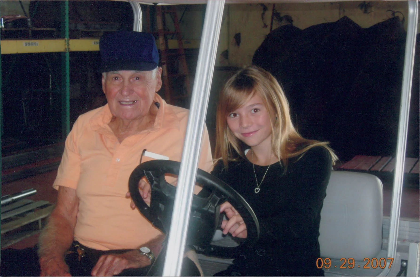 An elderly man and a young girl sitting in a golf cart. The man is wearing a navy hat and light orange shirt, and the girl has long blonde hair, a black shirt, and a necklace. The photo date is September 29, 2007.