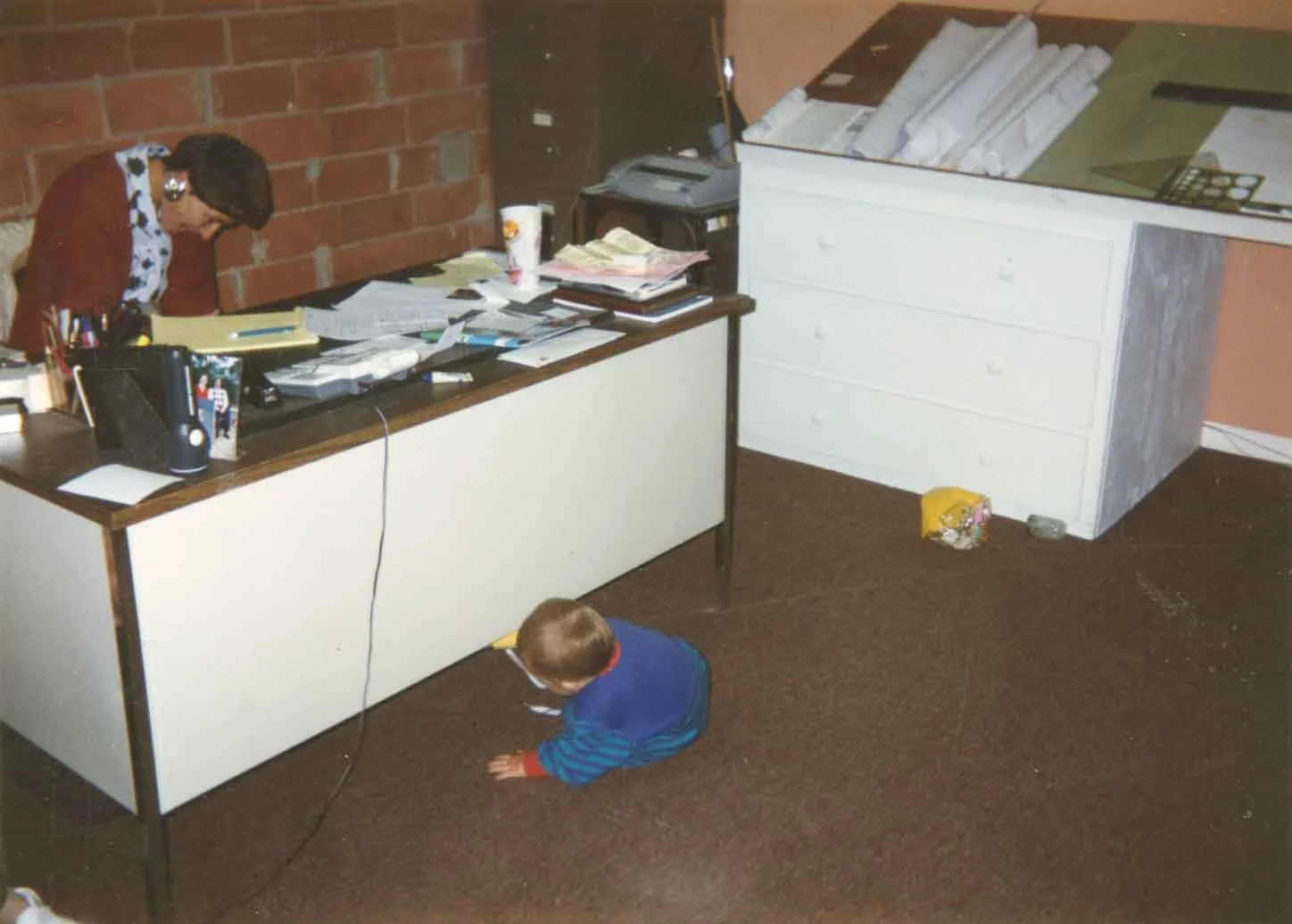 A cluttered office with a woman at the desk, a child on the floor, and various papers and objects scattered around.