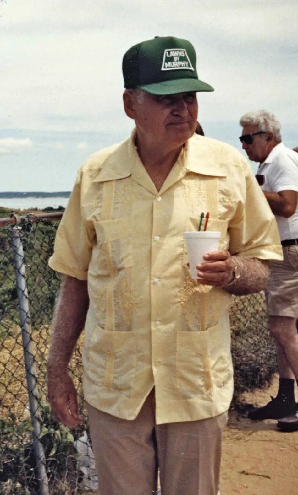 An older man wearing a yellow short-sleeve shirt, beige pants, and a green baseball cap that says "Lawns by Murphy." He is holding a white styrofoam cup with two pens inside. There is a man in a white shirt and sunglasses in the background, standing