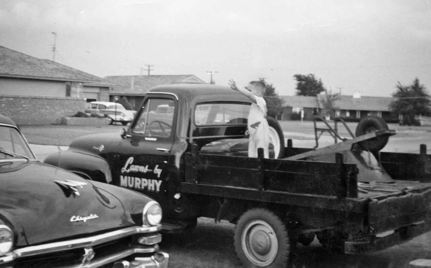 A young boy standing in the bed of a vintage pickup truck, waving. The truck is parked on a street with houses in the background, black and white photo.