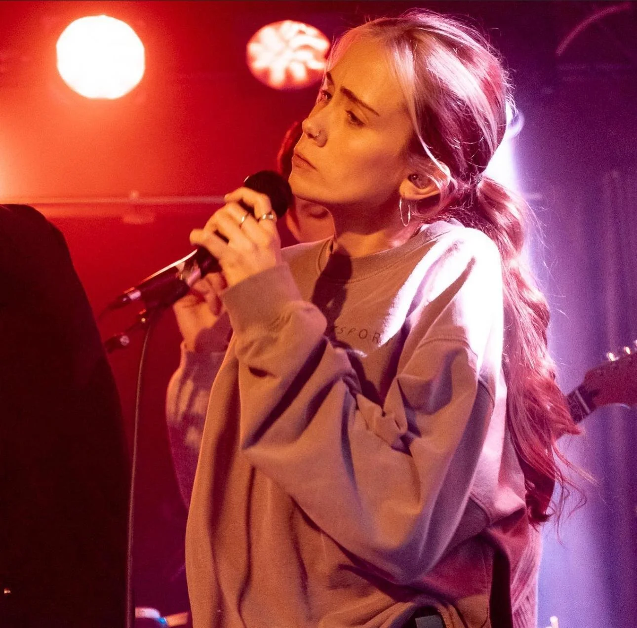 A woman with long, wavy hair holding a microphone on stage, illuminated by red and purple stage lights.