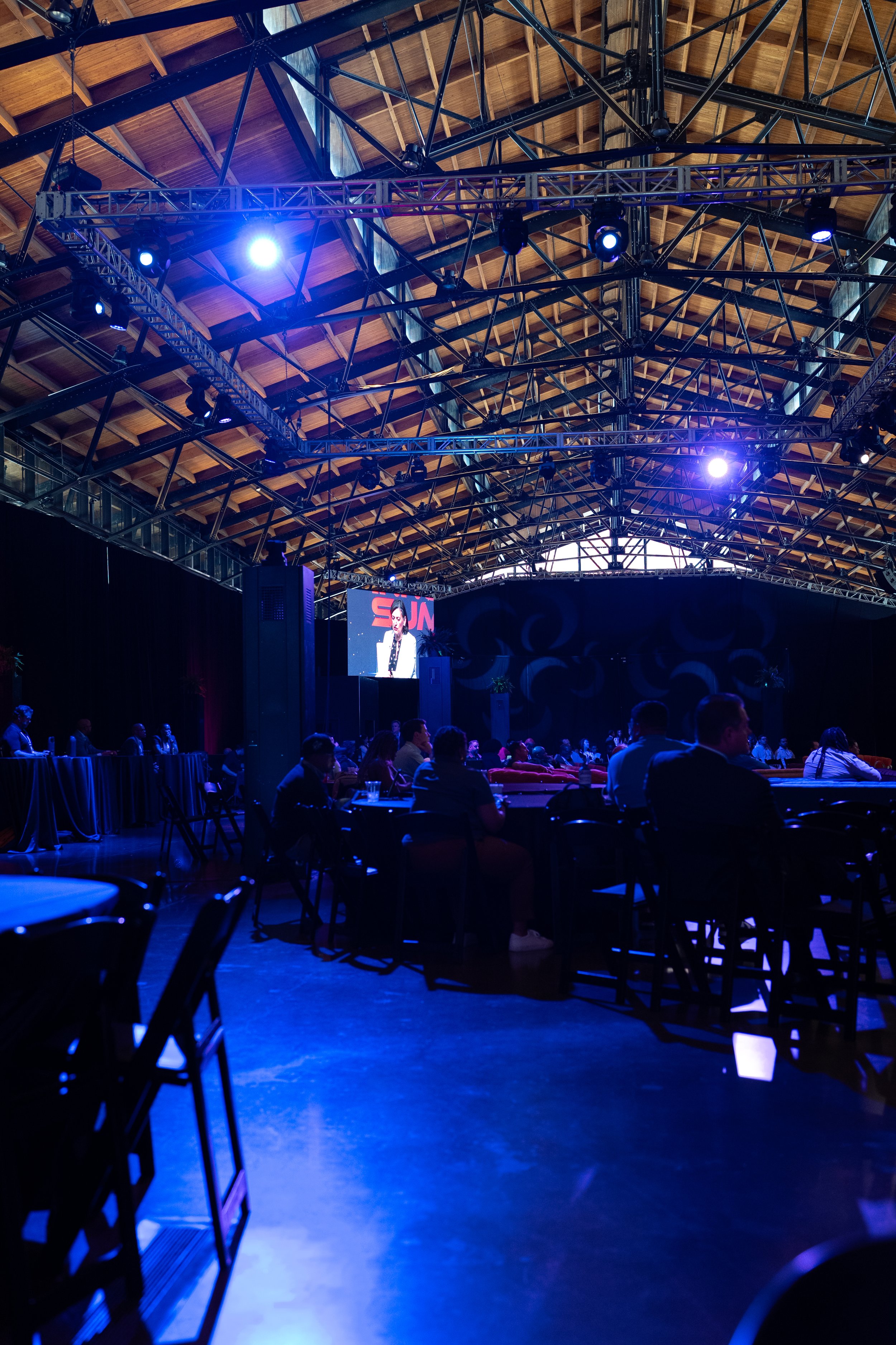 An indoor event space with a high wooden ceiling and metal framework, featuring an audience seated at tables facing a brightly lit stage with a large screen. The screen displays a person speaking at a podium.