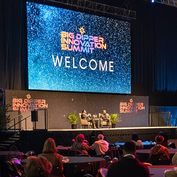 Large screen displaying 'Big Dipper Innovation Summit' with a starry background and 'Welcome' written underneath, stage with two people seated in conversation, audience seated facing the stage, decorated with potted plants.