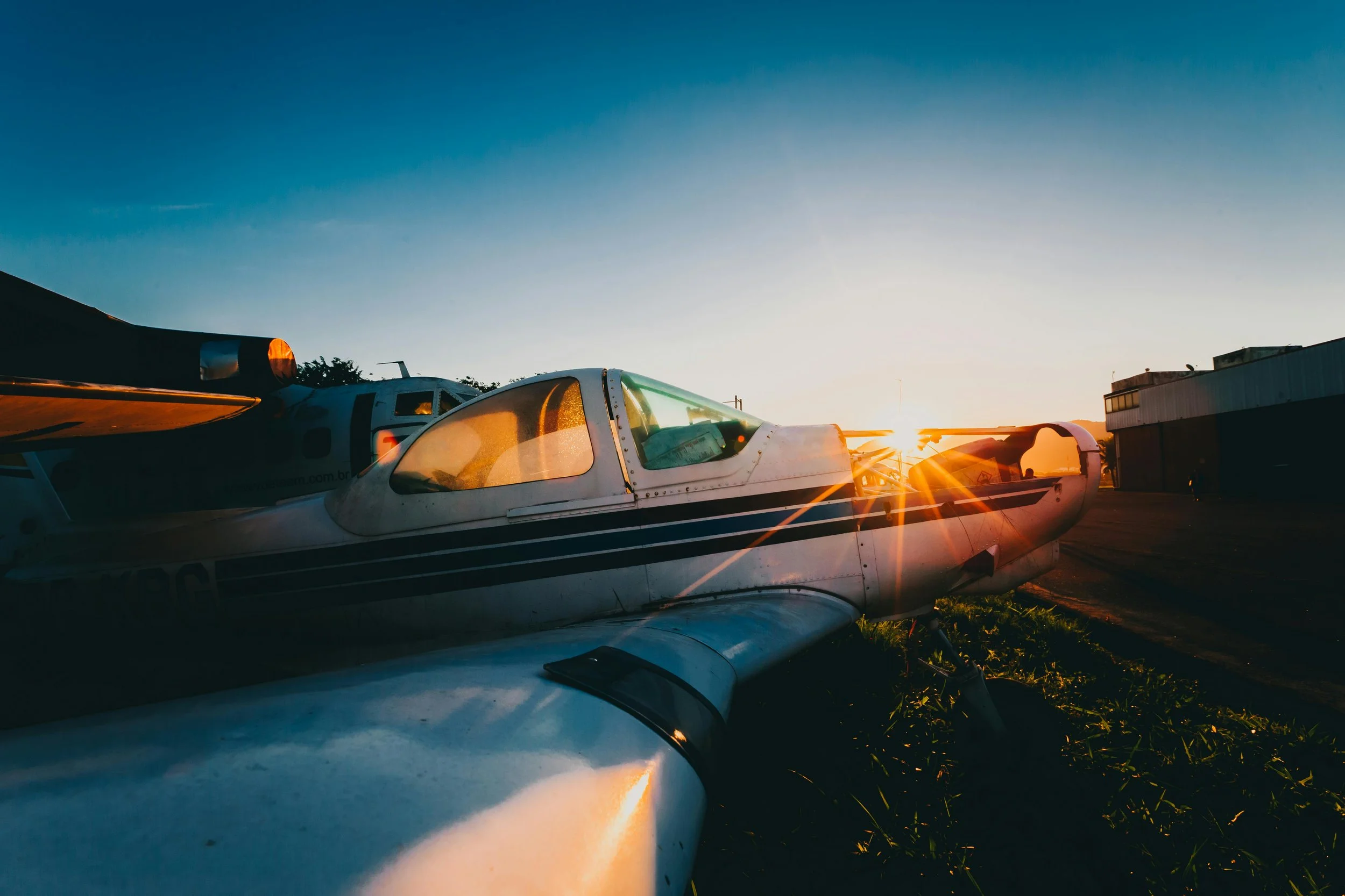 Small white airplane parked on the grass at sunset, with the sky clear and a building in the background.