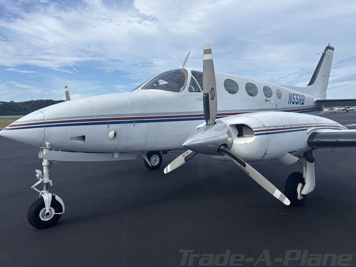 Small twin-engine propeller aircraft parked on tarmac, with weathered tarmac background and cloudy sky.