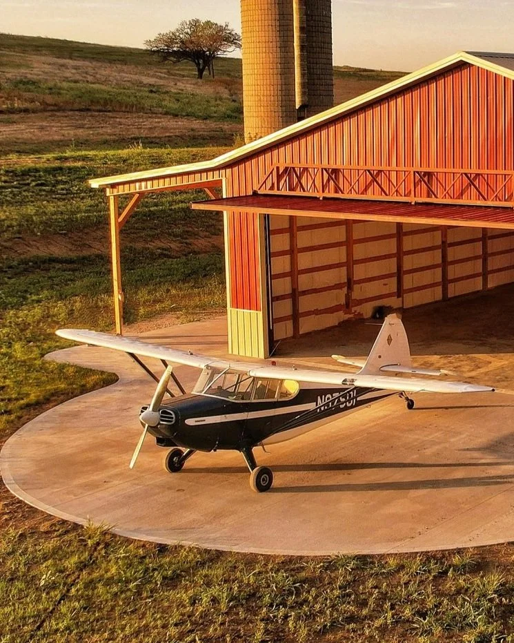 A small airplane parked outside a red barn near a silo on a farm, with a grassy Hill and a tree in the background during sunset.