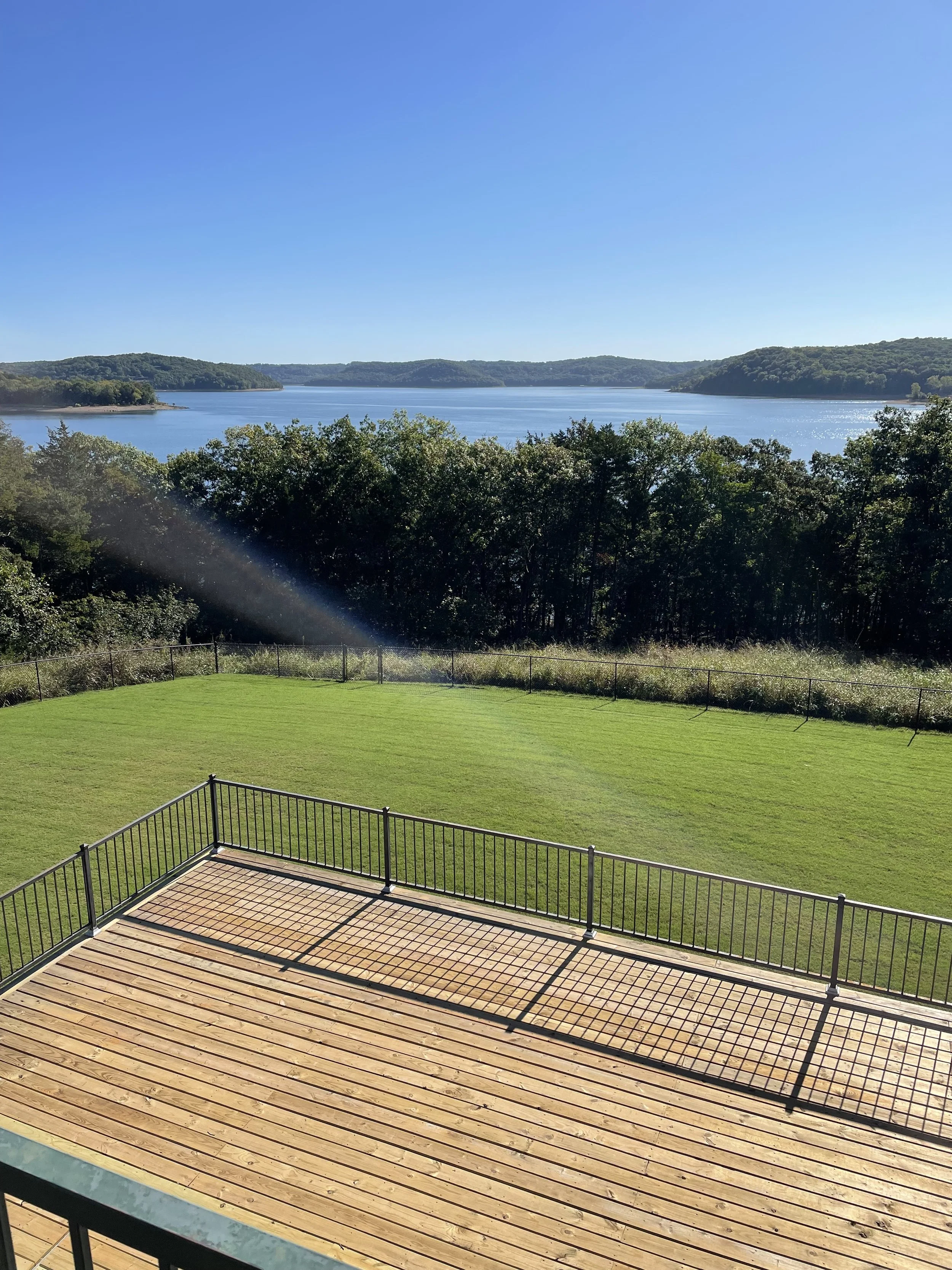 View of a lake with surrounding hills, a grassy yard, and a wooden deck with a black metal railing in the foreground.