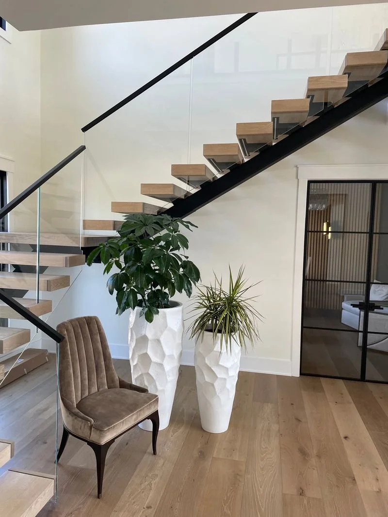 Indoor space with a modern staircase, two tall white textured planters with green plants, a vintage-style brown upholstered chair, and a black-framed glass door.