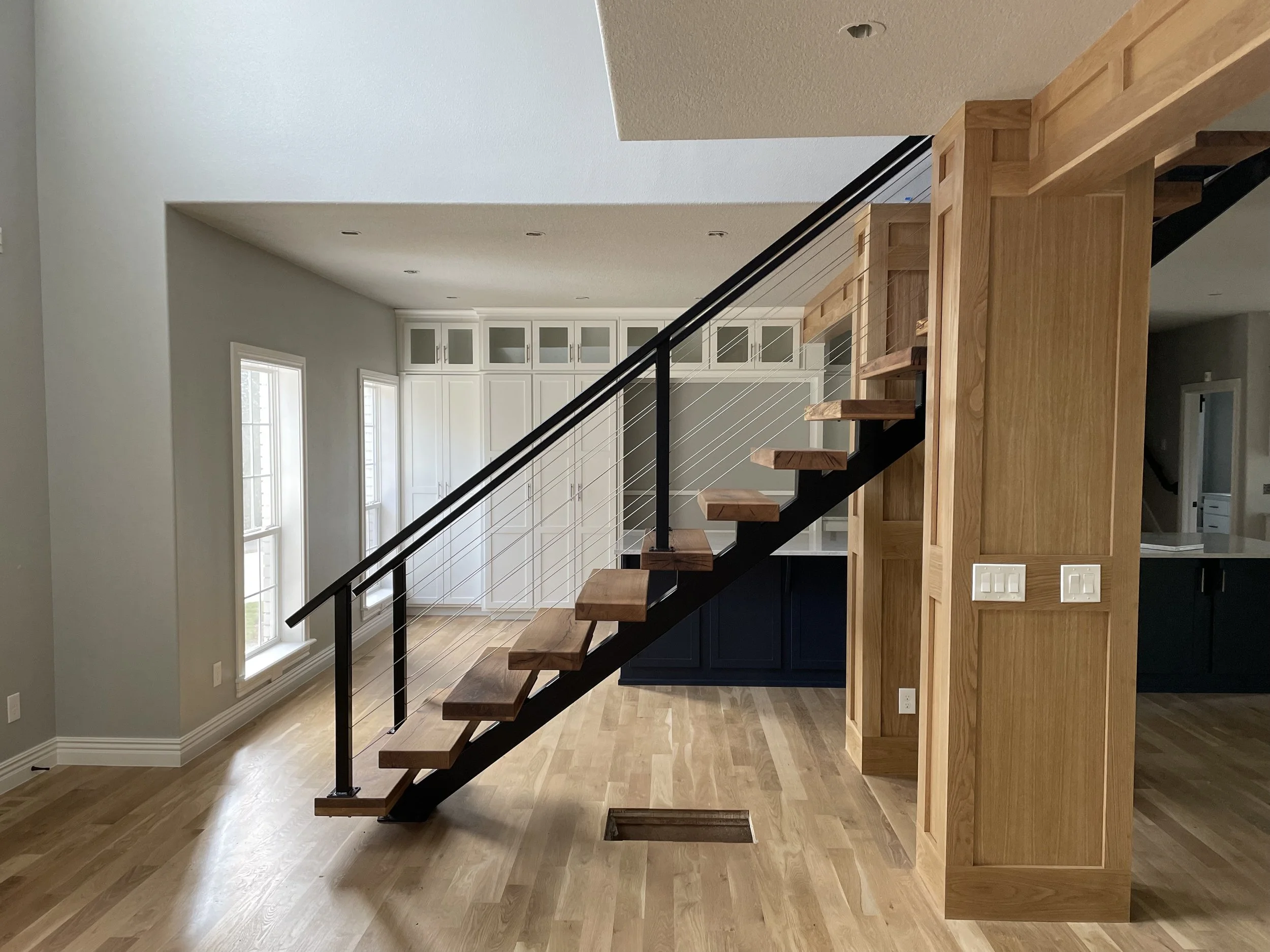 Interior of a modern home with a floating staircase made of wood and black metal, leading to the second floor. There are three windows on the left wall, a built-in cabinet with glass-fronted upper doors, and a mix of wood and painted cabinetry in the background. The floor is hardwood, and the walls are painted in light neutral colors.