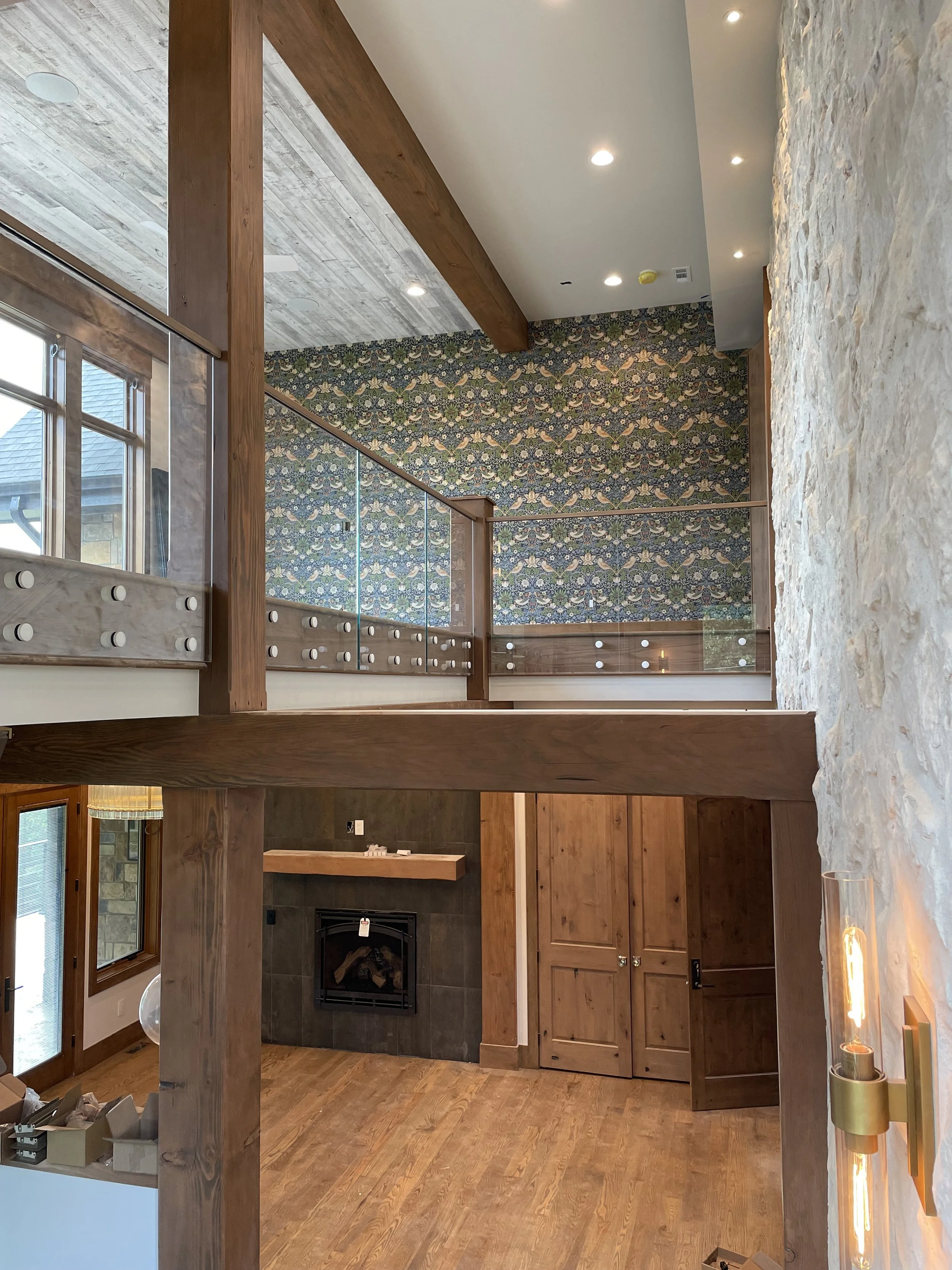 Interior view of a modern home with a second-floor balcony overlooking a living area with a stone fireplace and wooden cabinetry, featuring wood and stone finishes, patterned wallpaper, and recessed ceiling lights.