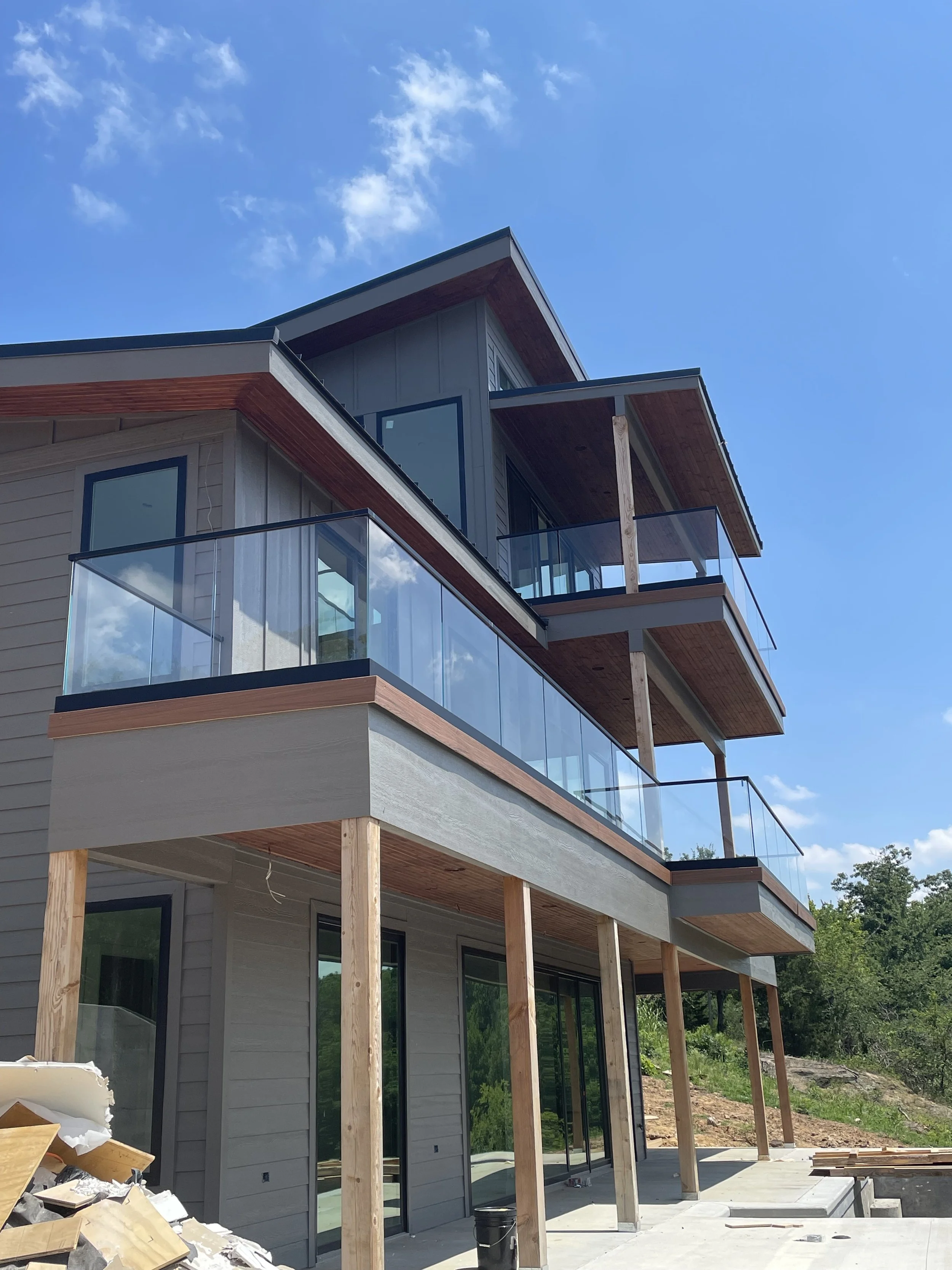 A modern multi-story house under construction with large glass windows and VIEWRAIL balconies, supported by wooden beams, against a blue sky with clouds.