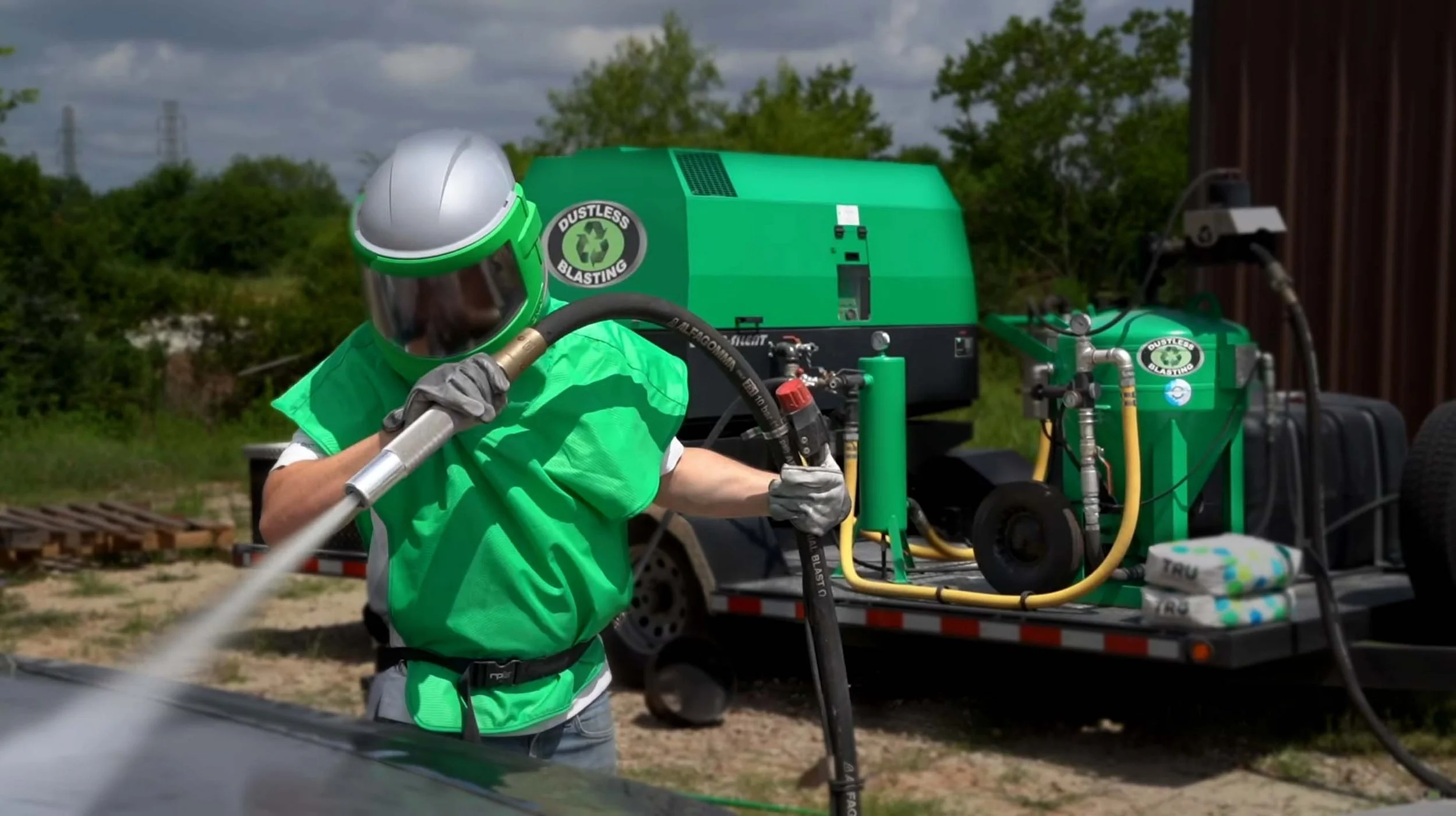 A person in protective gear using Dustless Blasting to clean a surface outdoors, with green equipment labeled 'Dustless Blasting' in the background.