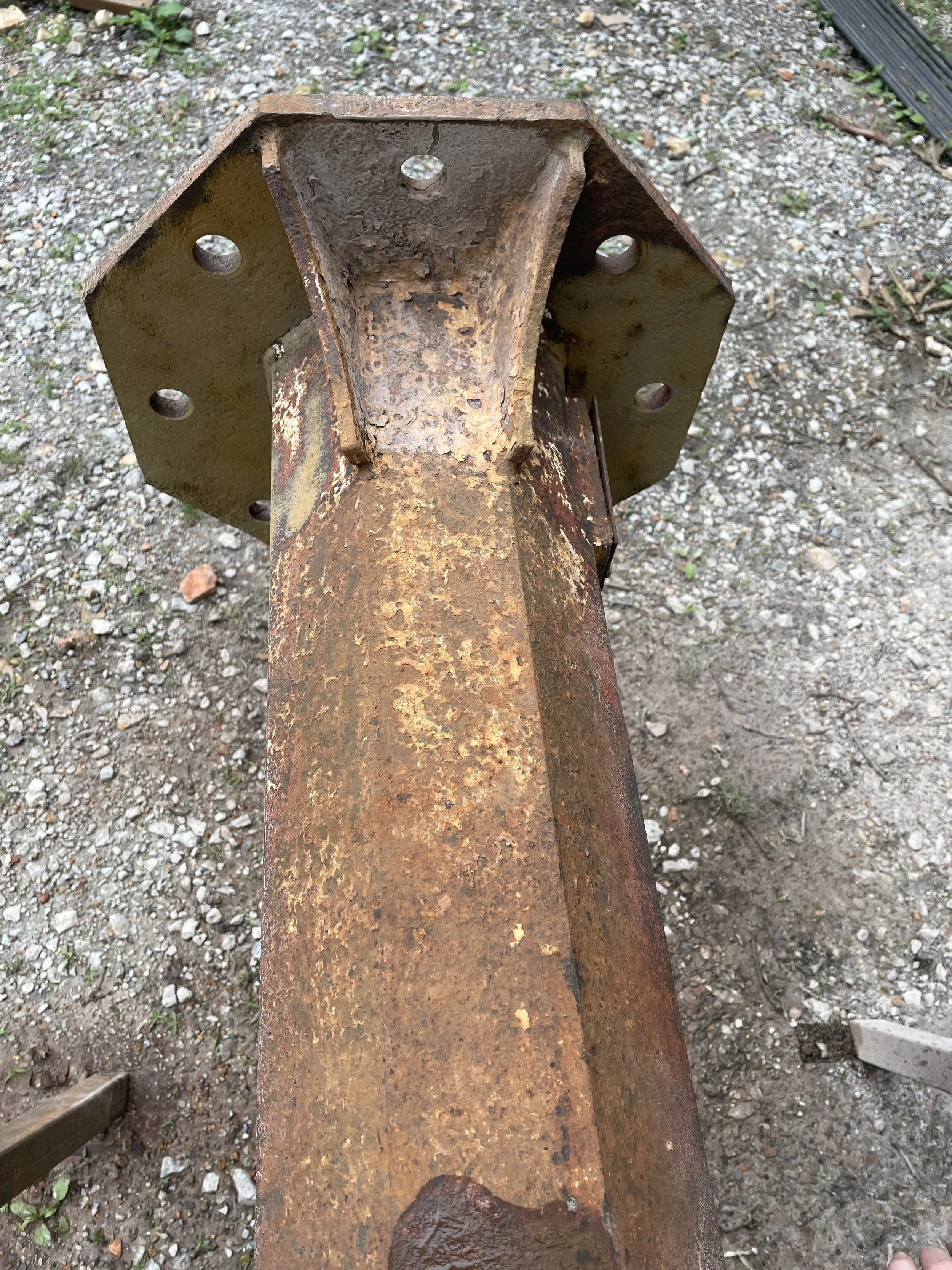 Rusty metal beam with a seat mounting bracket on top, outdoors on a gravel surface.