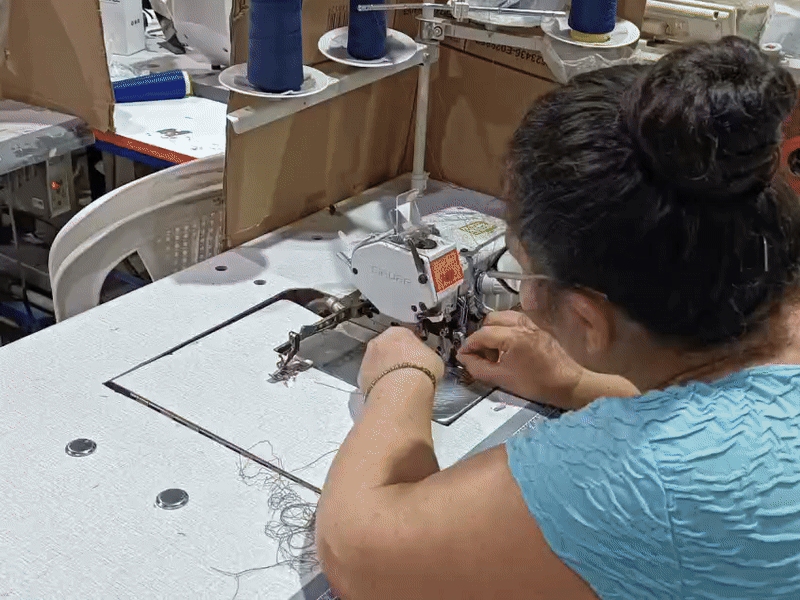 Una mujer trabajando en una máquina de coser industrial en un taller de fabricación, con bobinas de hilo azul en el fondo.