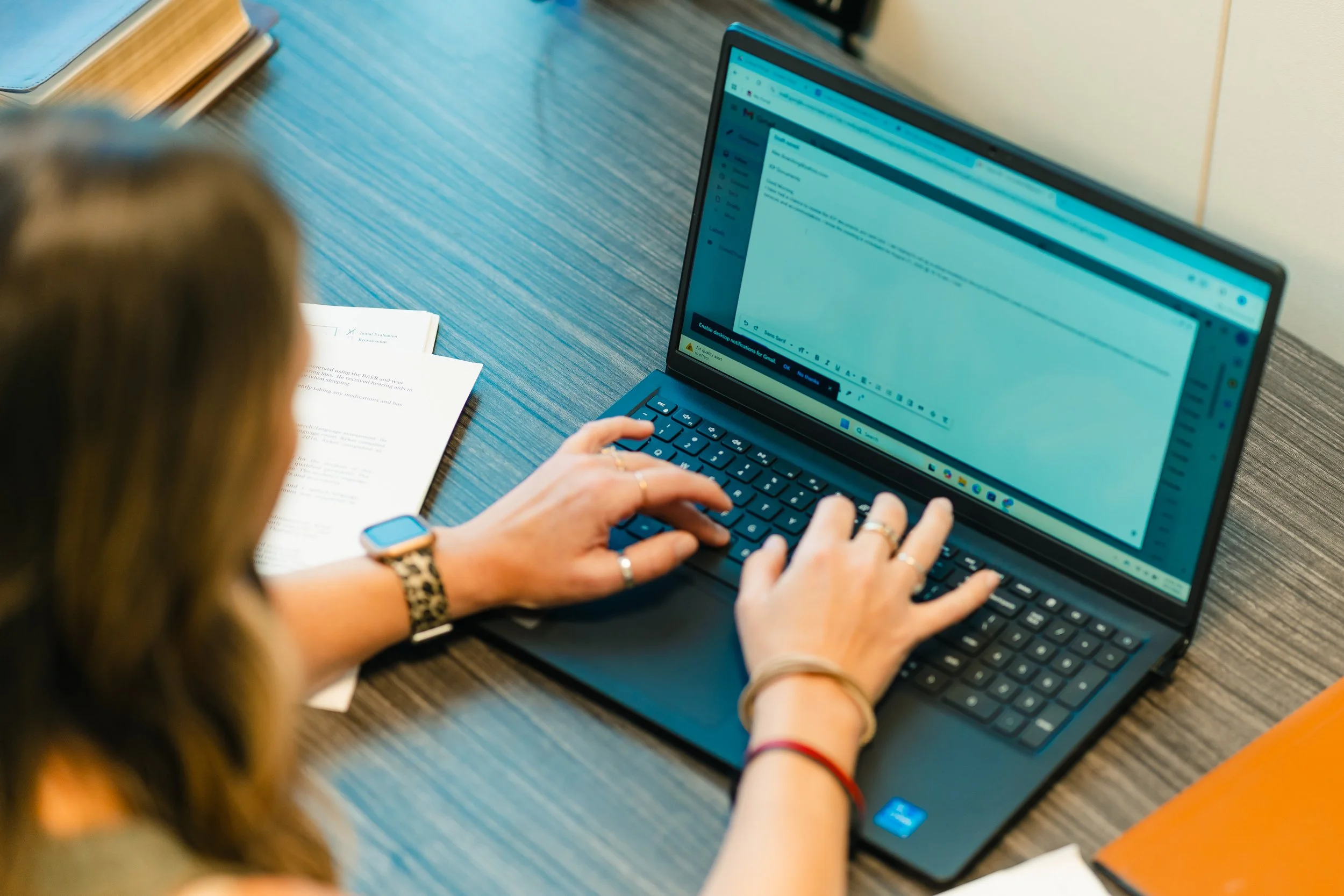 A woman with wavy hair, wearing a leopard print watch, working on a laptop at a wooden desk with papers and a closed notebook.