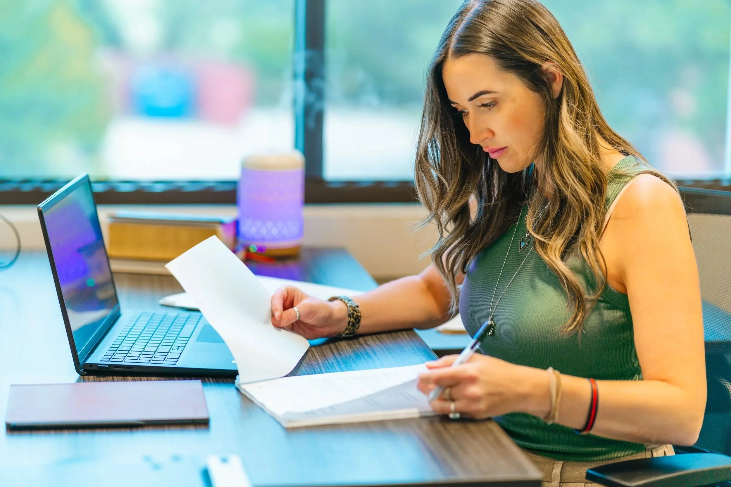 Woman in green sleeveless top working at desk with laptop, papers, and pen, near window with colorful blurred background.