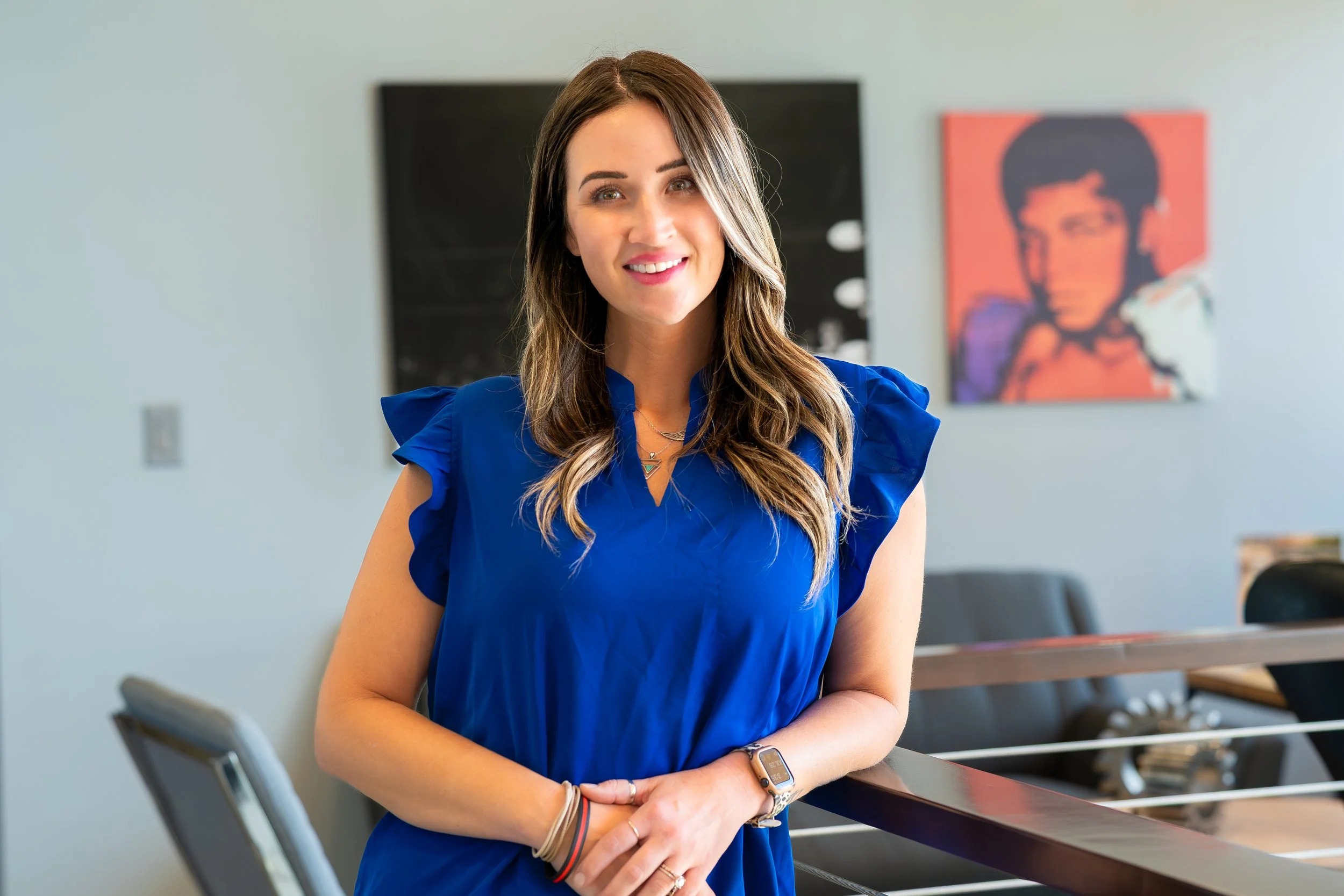A woman with long brown hair wearing a blue dress standing in a modern office setting.